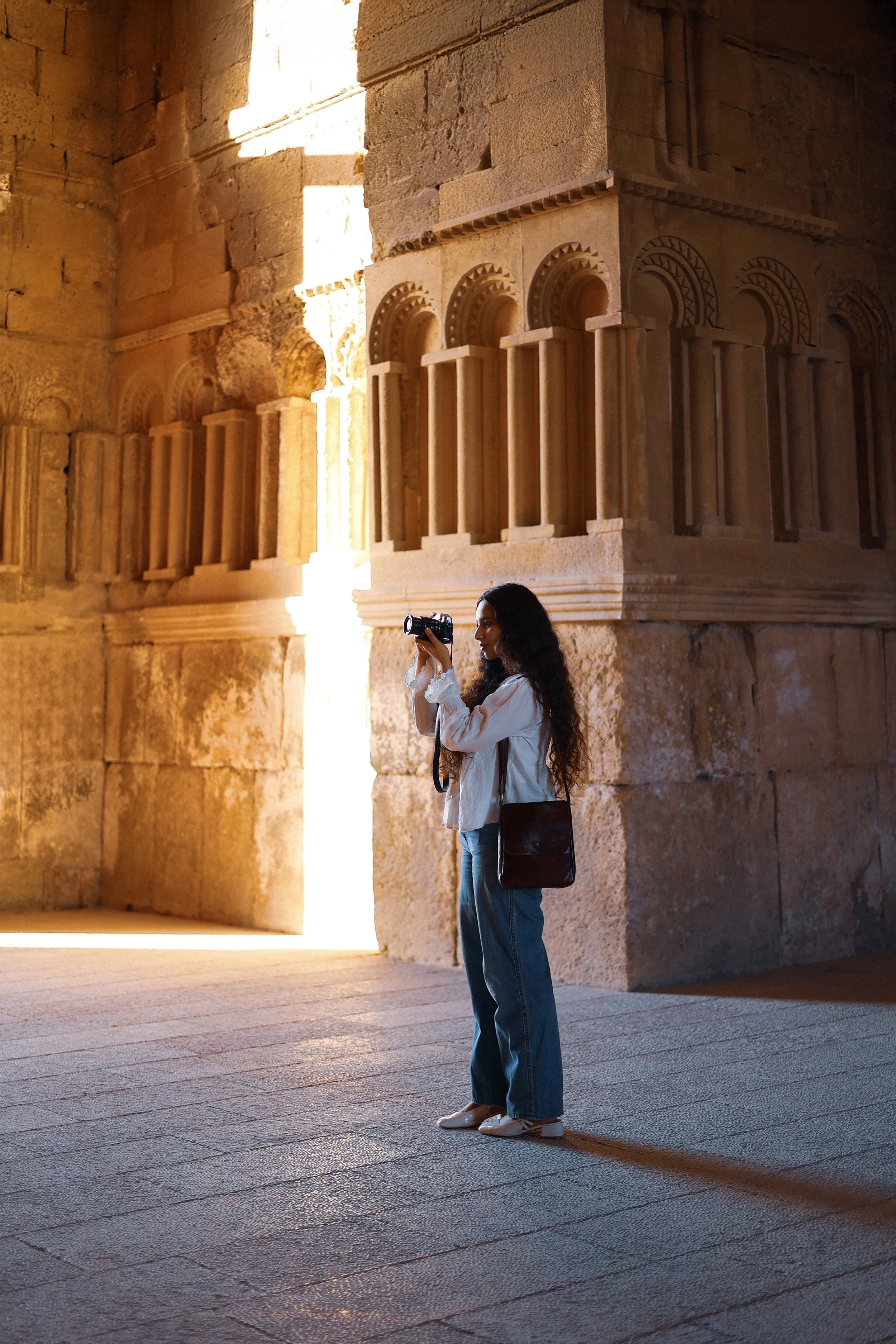 A woman with long dark hair, wearing a white shirt, blue jeans, and white shoes, stands inside an ancient stone structure, holding a camera and taking a photo. The structure features detailed stone carvings and archways, with sunlight streaming through an opening.