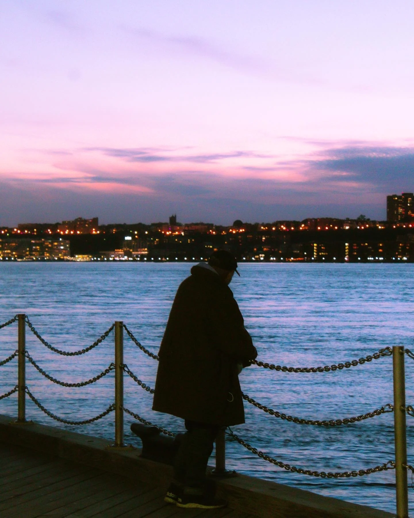 A person standing on a waterfront dock at sunset, looking out over the water with a city skyline in the distance and colorful sky above.