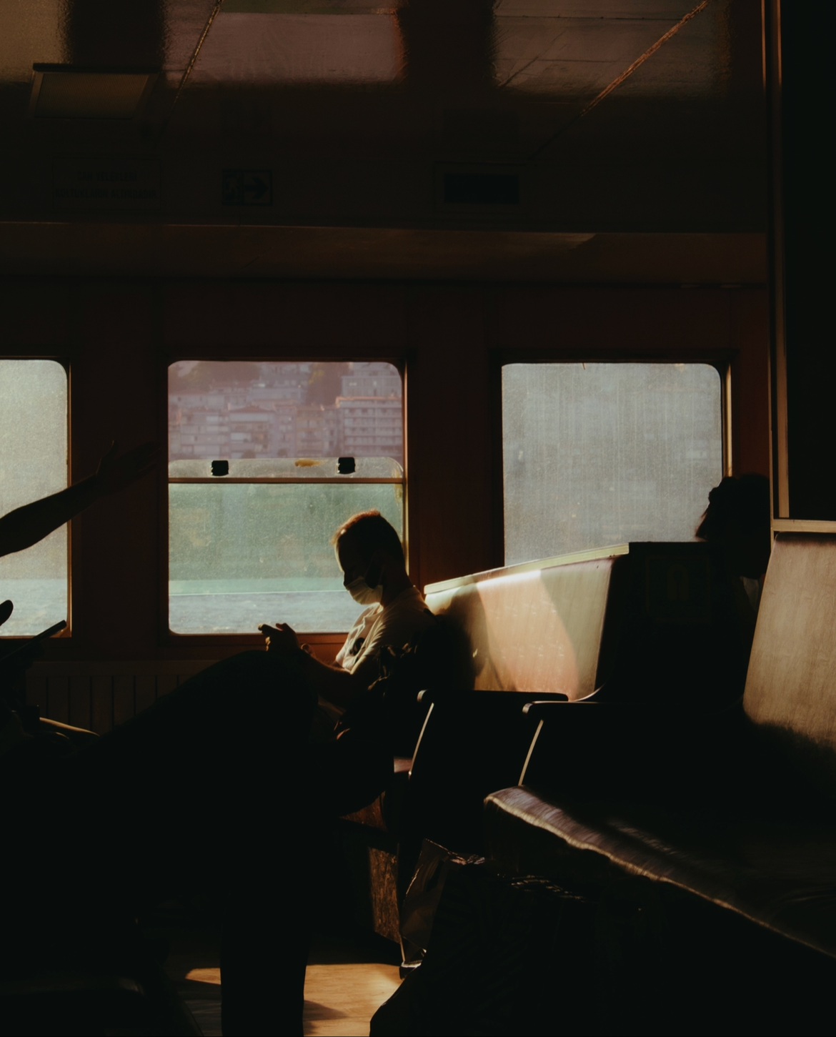 Inside a ferry or bus with people sitting and wearing face masks, backlit by sunlight coming through windows showing a cityscape outside.