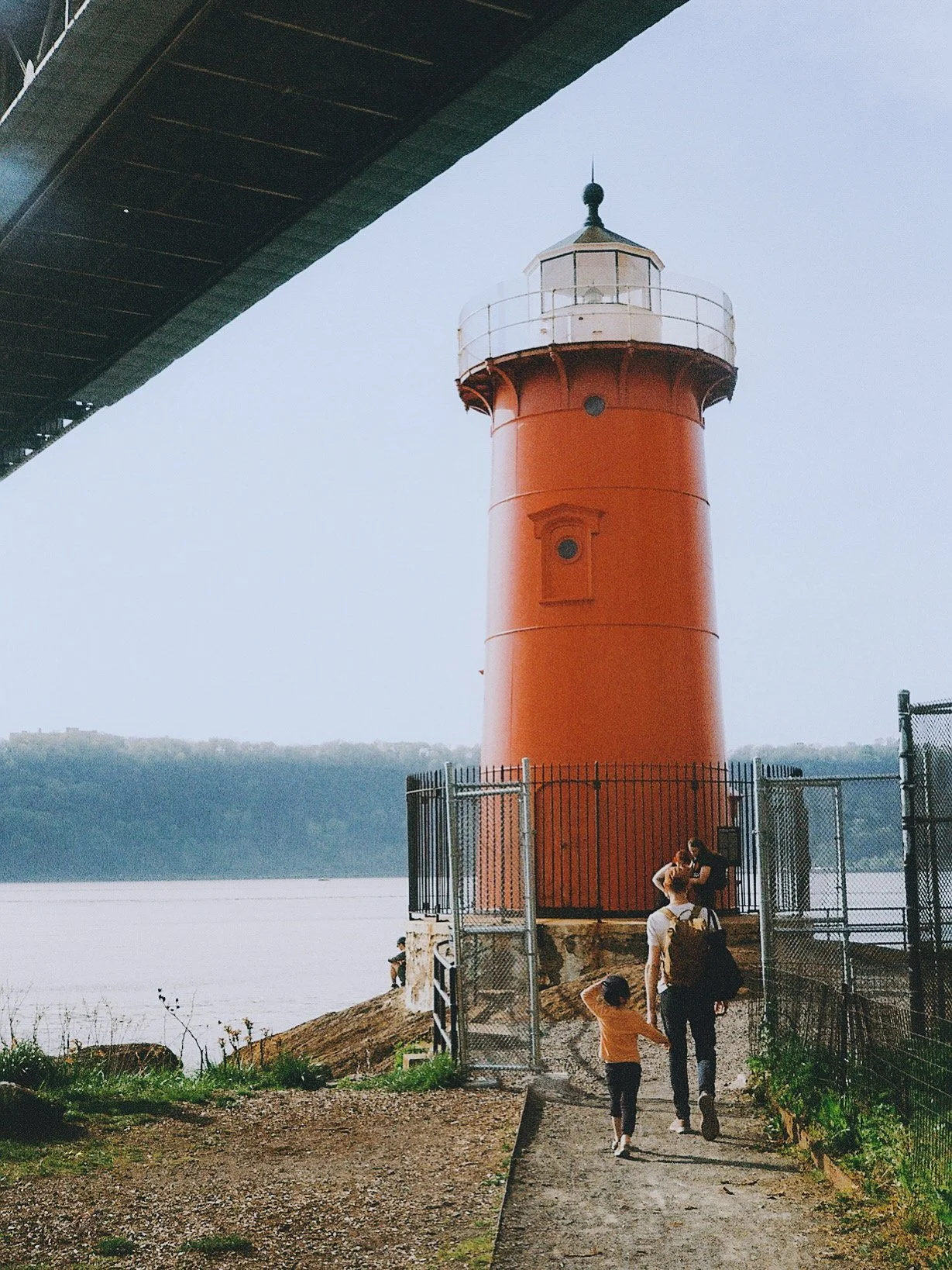 People walking towards a red lighthouse near a body of water, with a bridge overhead and a hilly landscape in the background.