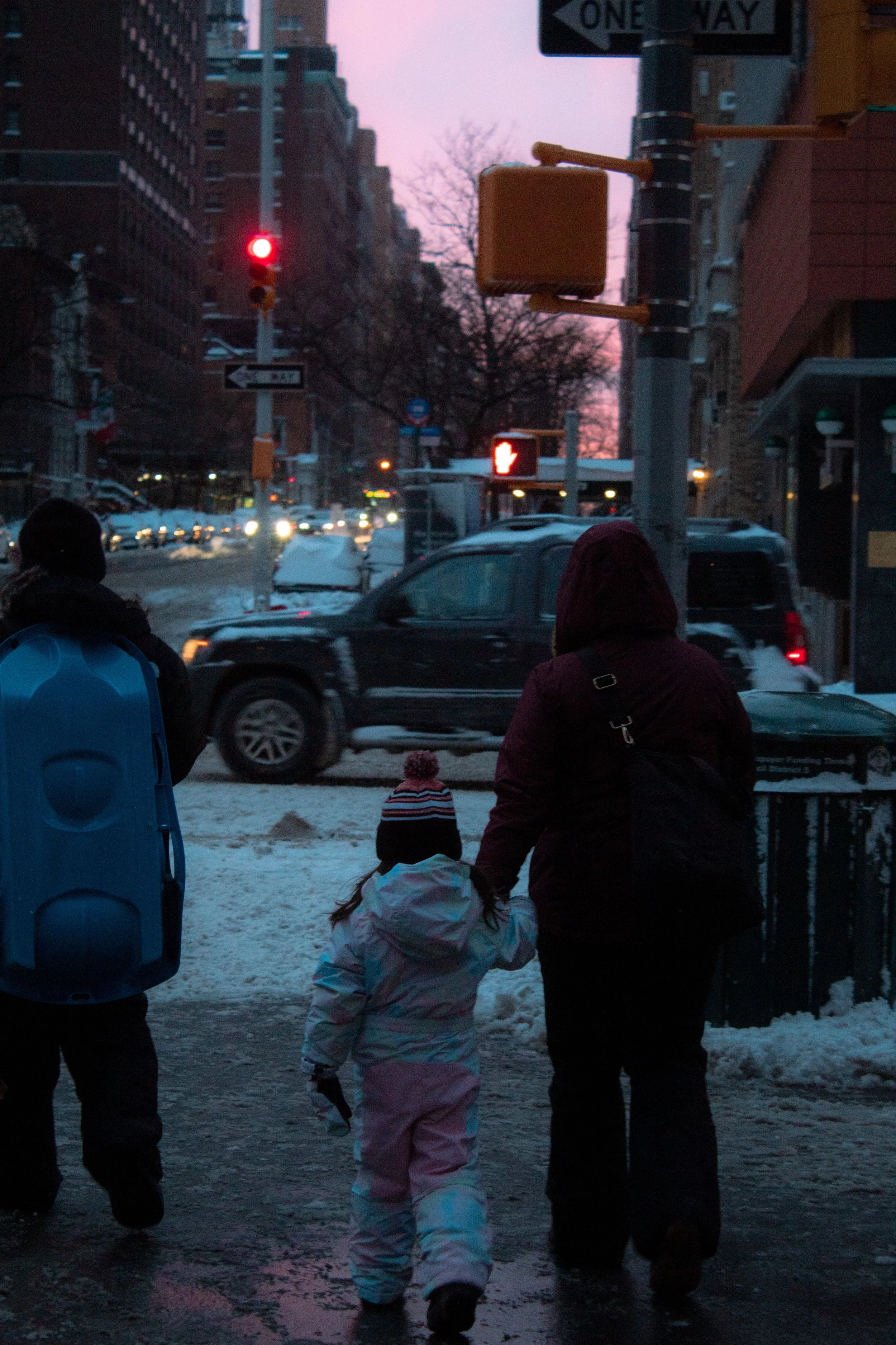 A street scene at dusk with a crosswalk and traffic lights. A woman and child in winter clothing walk hand in hand on the sidewalk, with snow on the ground. Cars and buildings are visible in the background.