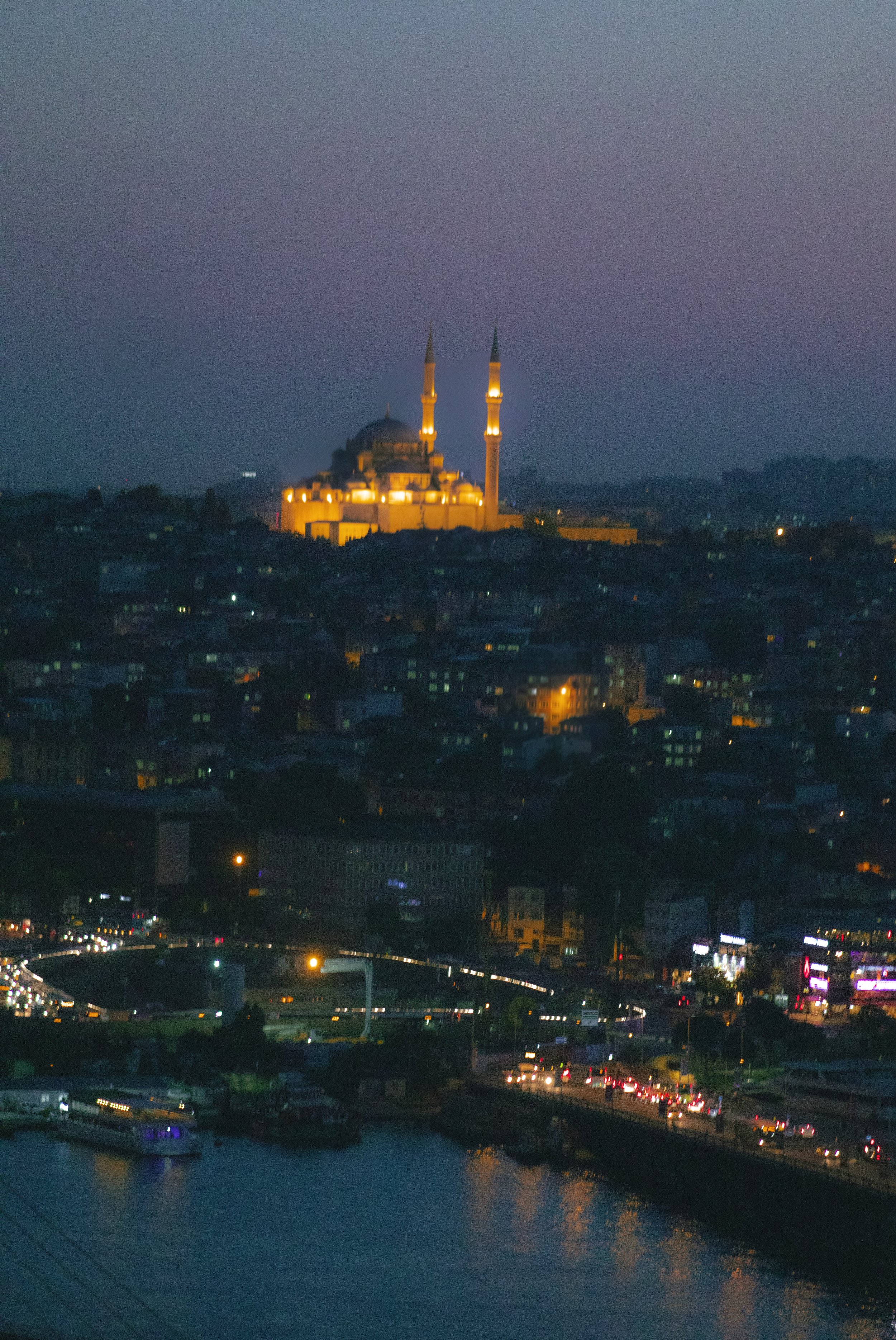 Nighttime cityscape of Istanbul, Turkey, featuring the illuminated Sultan Ahmed Mosque (Blue Mosque) with its domes and minarets visible, over a densely built urban area with streetlights and traffic, and a body of water in the foreground.