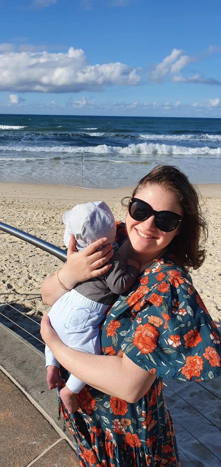 A woman wearing sunglasses and a floral dress holding a baby on a beach with ocean waves and a blue sky with clouds in the background.