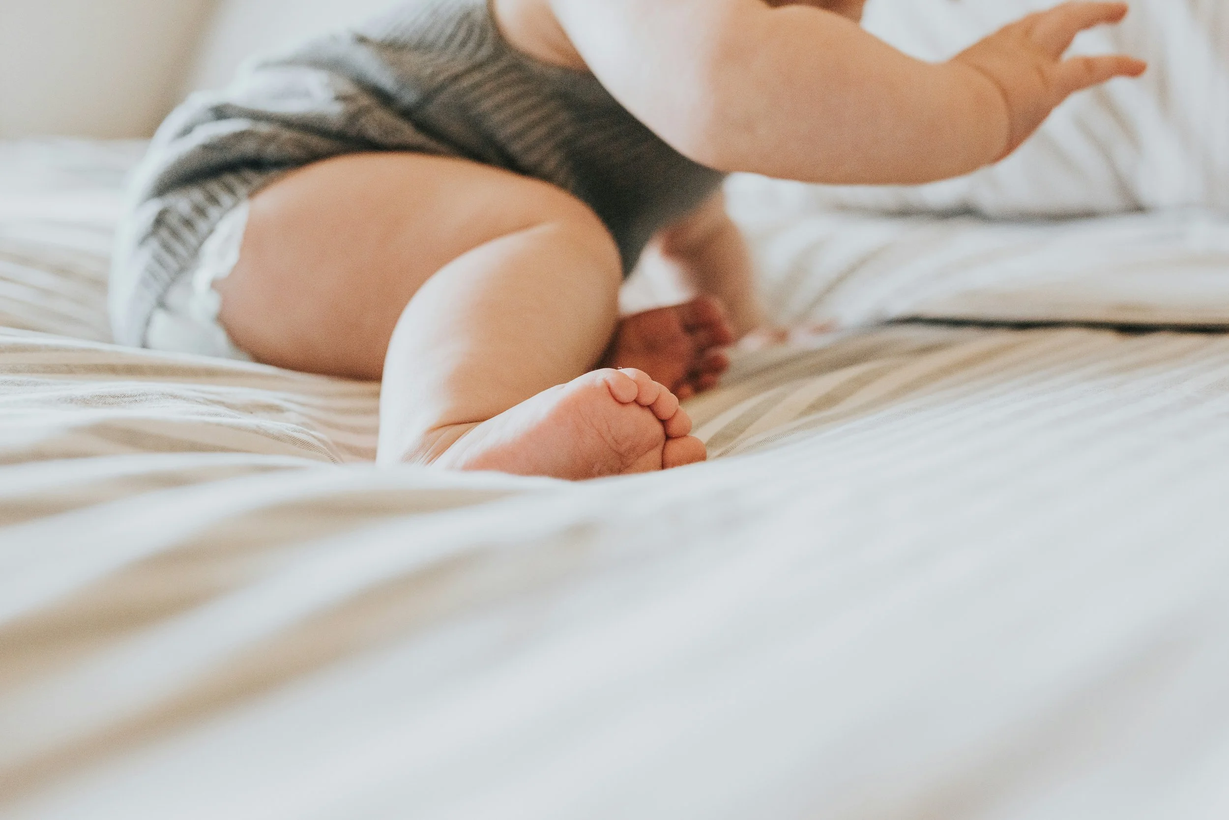 Close-up of a toddler crawling on a bed, wearing a striped shirt and diaper, with part of their face and hand visible.