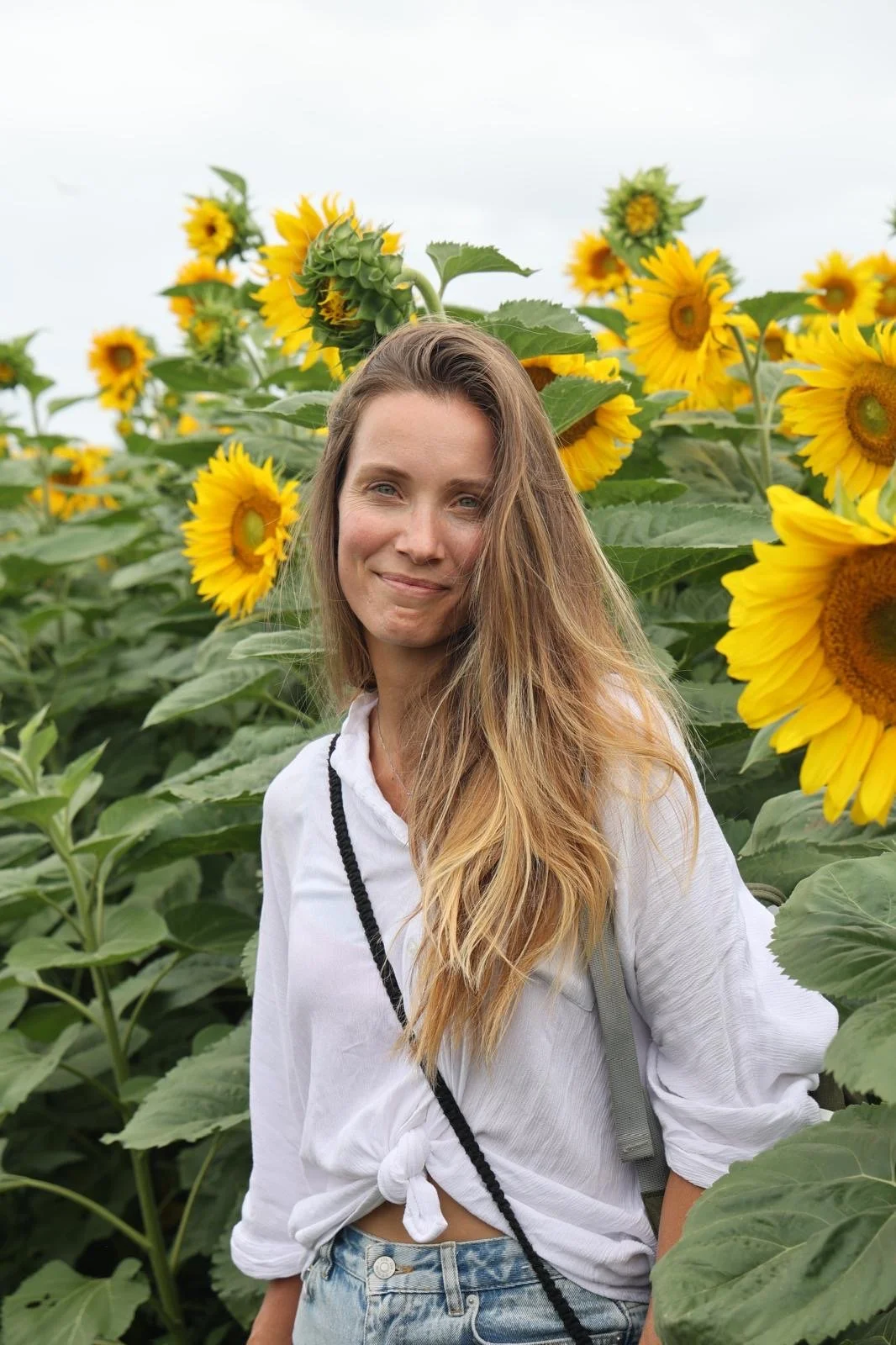 A woman with long blonde hair standing in a sunflower field, smiling at the camera.