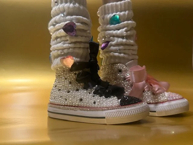 A pair of designer sneakers decorated with pearls, rhinestones, and large bows, along with plush socks adorned with colorful heart-shaped embellishments and rhinestones.