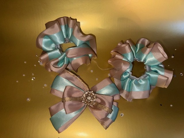 Two decorative bows made of metallic gold and teal ribbons, attached with a small rhinestone brooch, placed on a reflective gold surface.