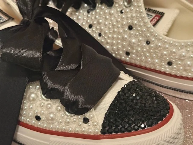 Close-up of decorated sneakers with pearl and black rhinestone embellishments, black ribbon laces, and white soles with red accents.