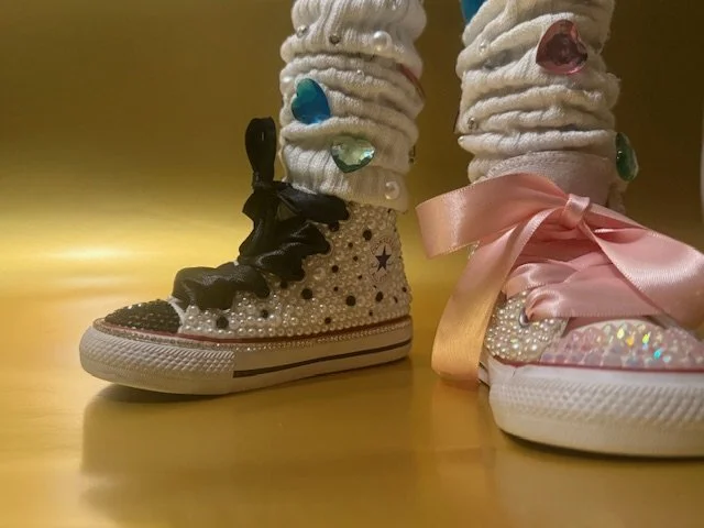 Close-up of two decorated sneakers with pearl embellishments and fabric bows, one with black laces and the other with pink satin ribbon, on a yellow surface.