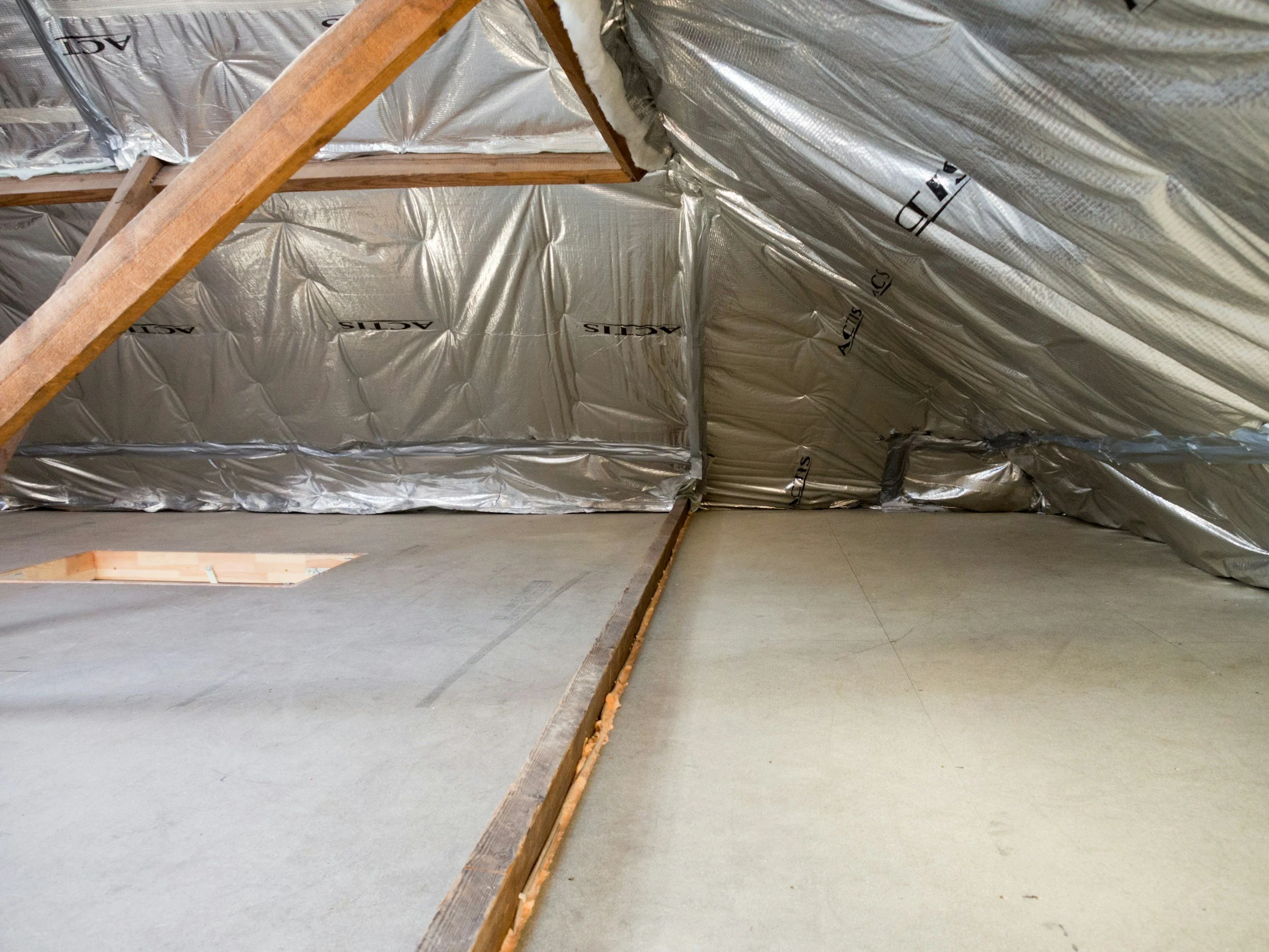 View of an attic space with insulation and wooden beams, unfinished ceiling with a narrow opening in the floor or roof.