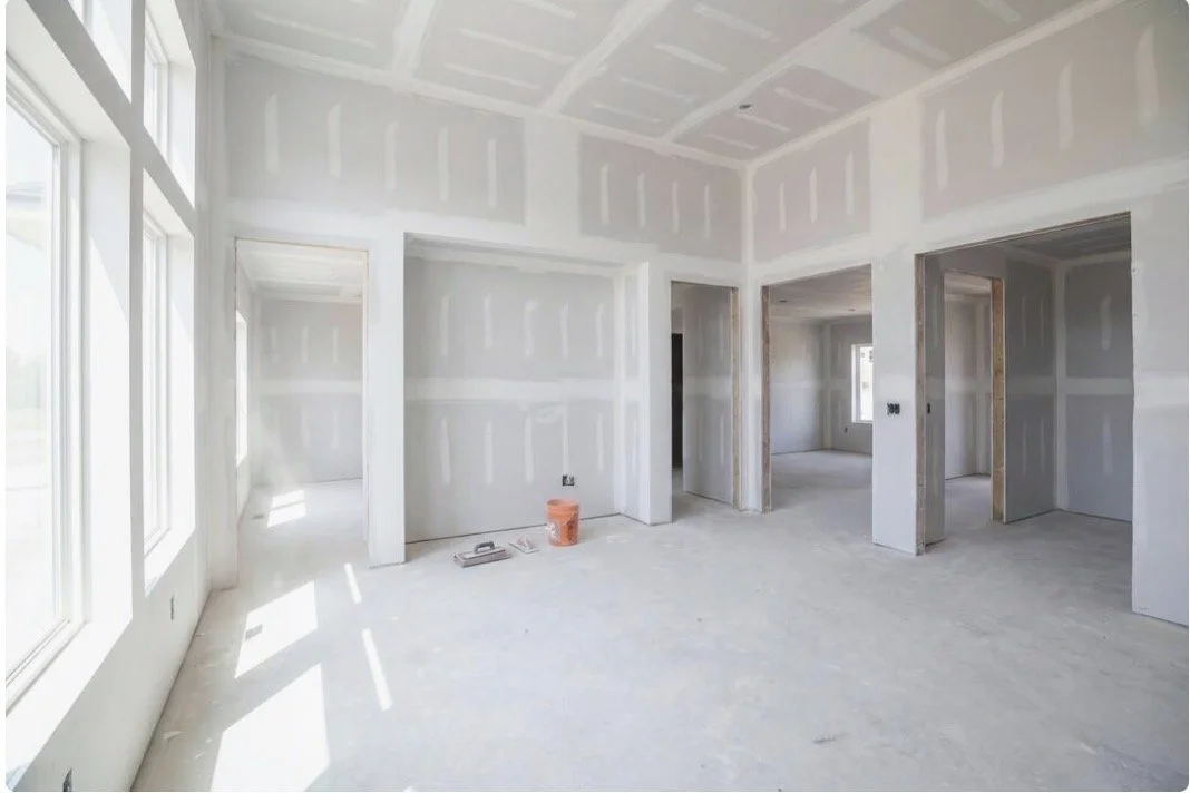 An unfinished room in a house under construction, with drywall walls and ceiling, large windows, and empty door frames.