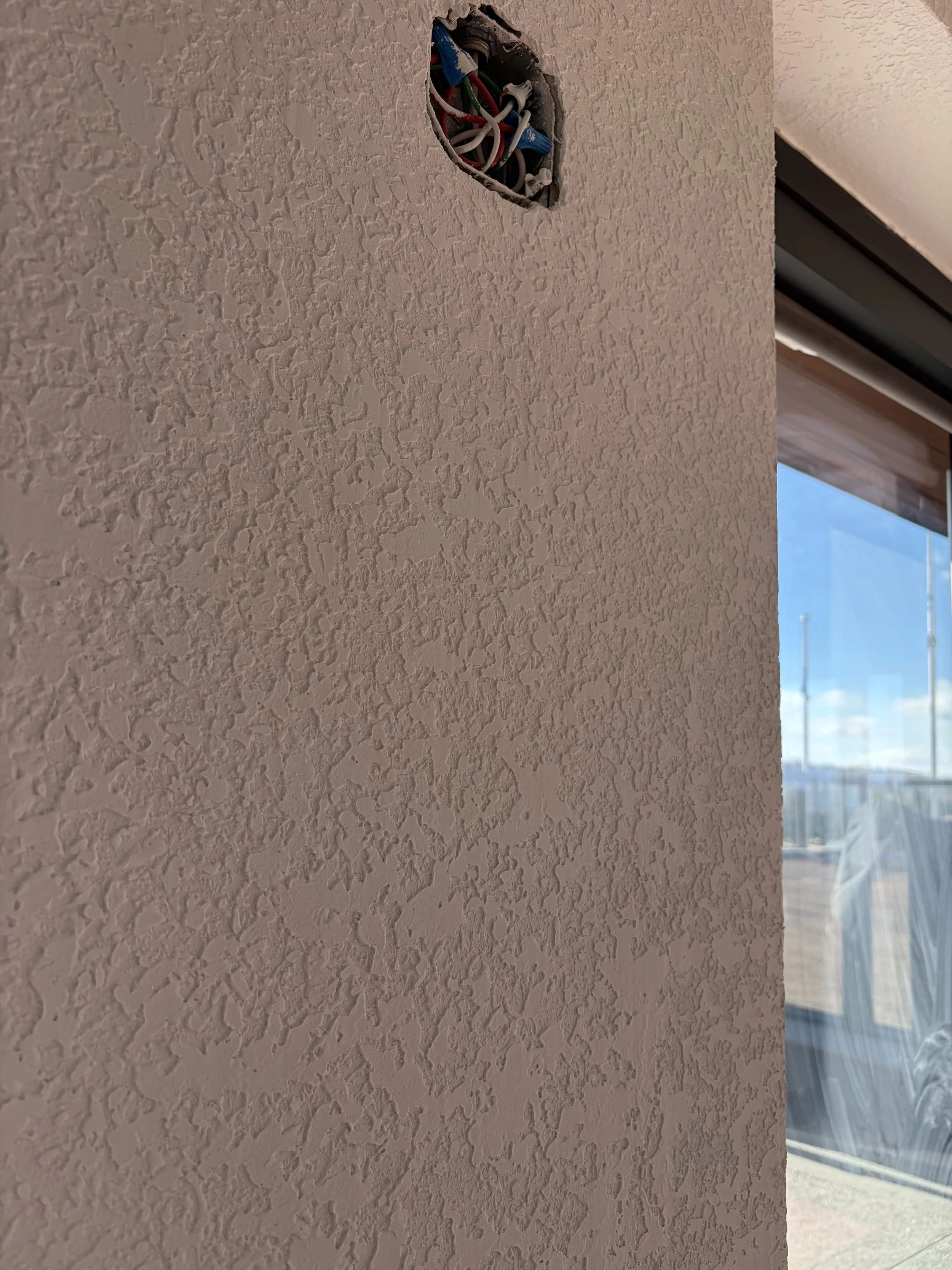 Ceiling with an open electrical box containing wires and a textured pink wall, with a window and outside view in the background.