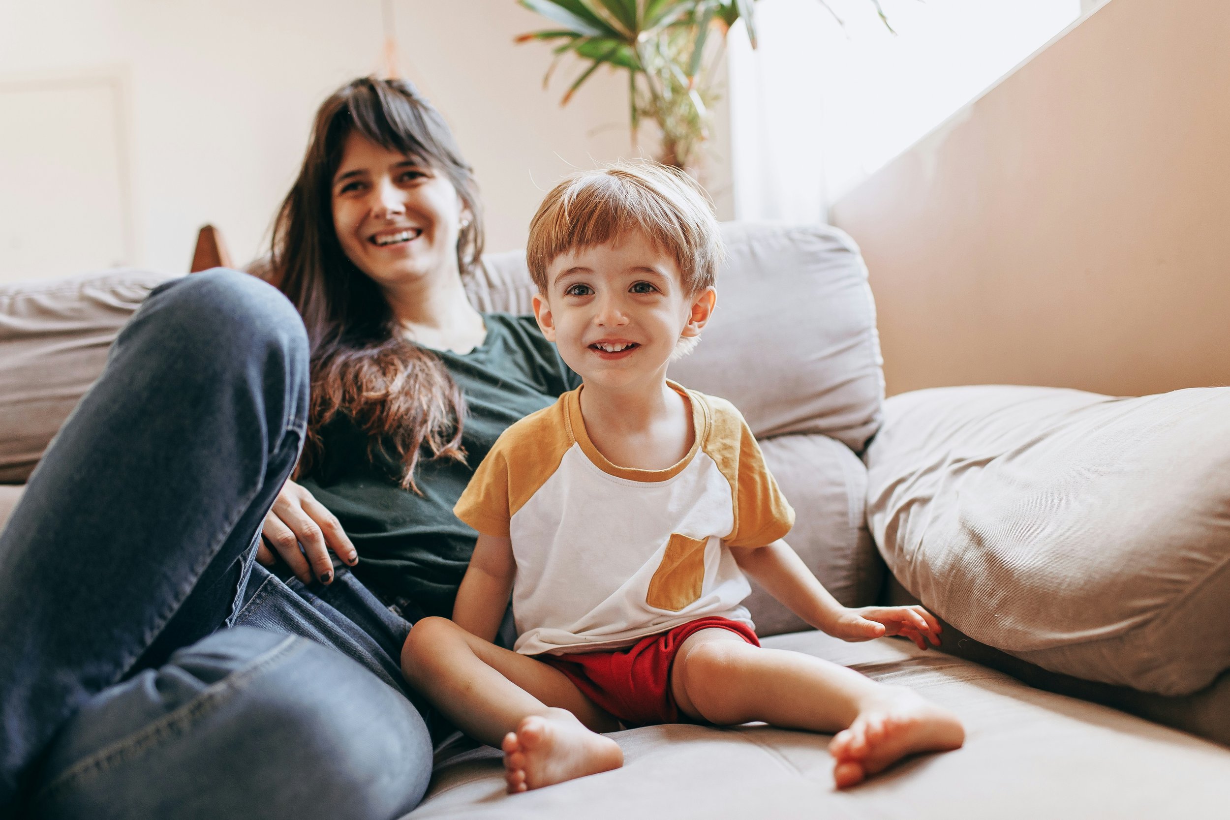 A smiling young woman and a happy young boy sitting on a beige couch in a well-lit living room. The woman has long brown hair and is wearing a dark green shirt and jeans. The boy has short light brown hair, a white and yellow shirt, and red shorts. A potted plant is visible in the background near the window.