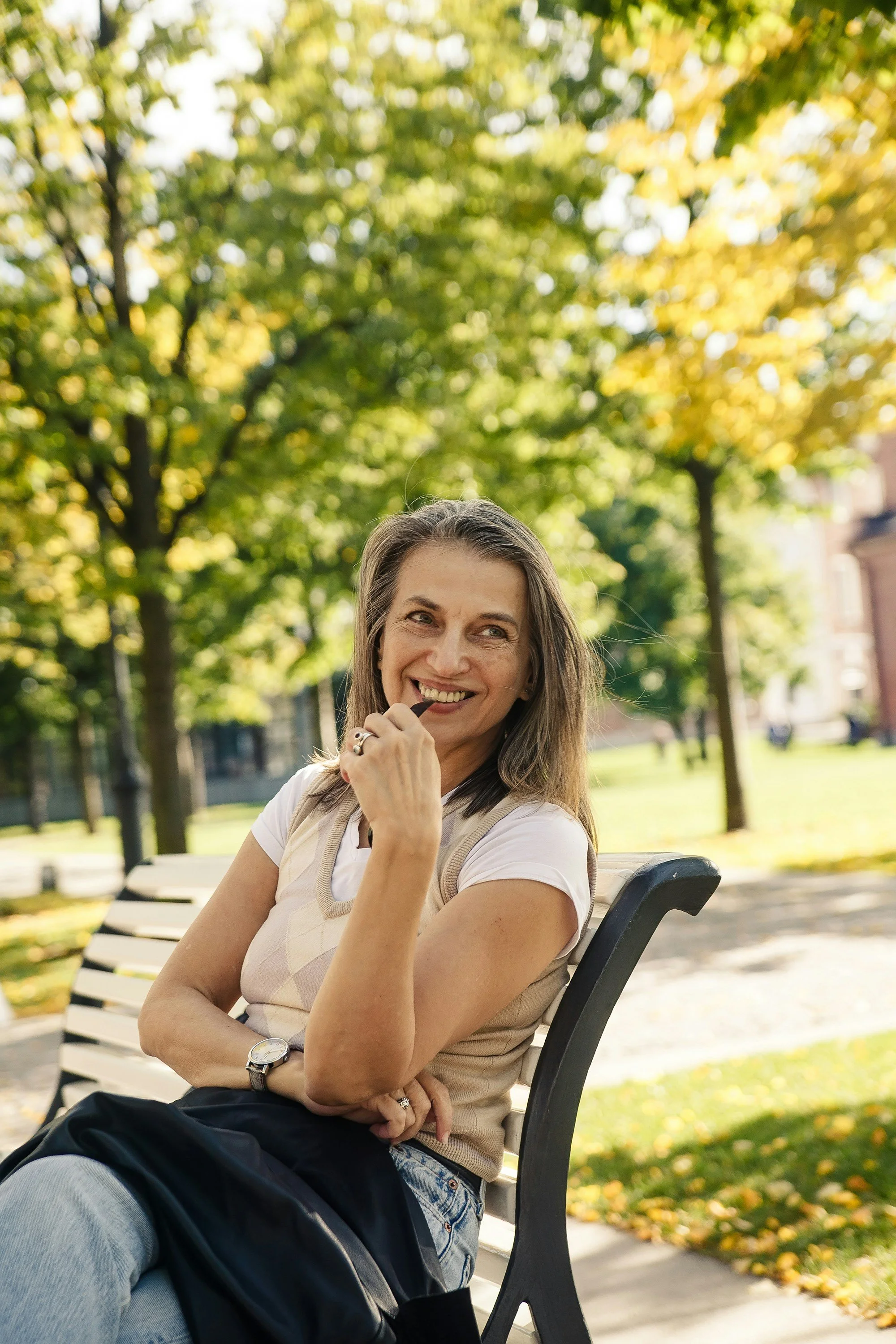 A woman sitting on a park bench, smiling and tilting her head with her hand near her mouth. She has long hair and wears a white t-shirt, beige vest, and a watch. The background shows trees with green and yellow leaves, indicating autumn.