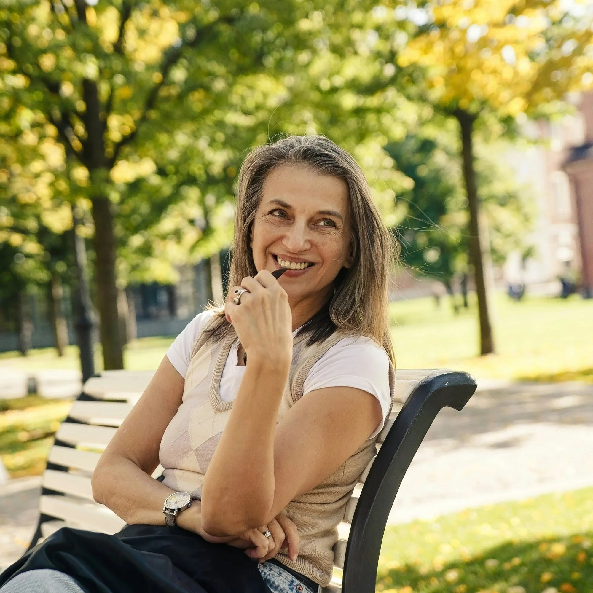 A woman sitting on a park bench smiling, with green trees and sunlight in the background.