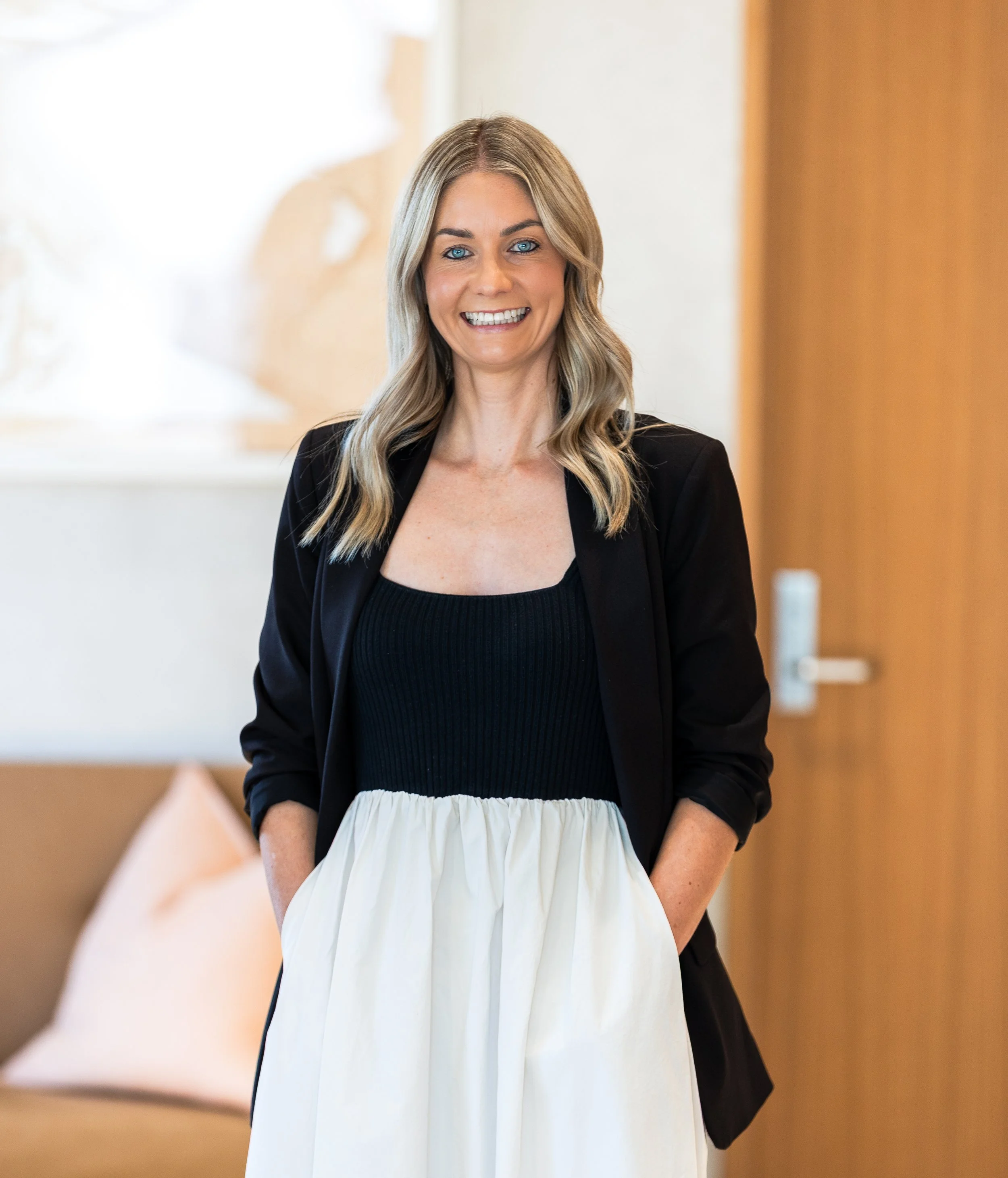 A woman with blonde hair and blue eyes smiling, wearing a black top, black blazer, and a high-waisted white skirt, standing indoors near a door with a blurred background.