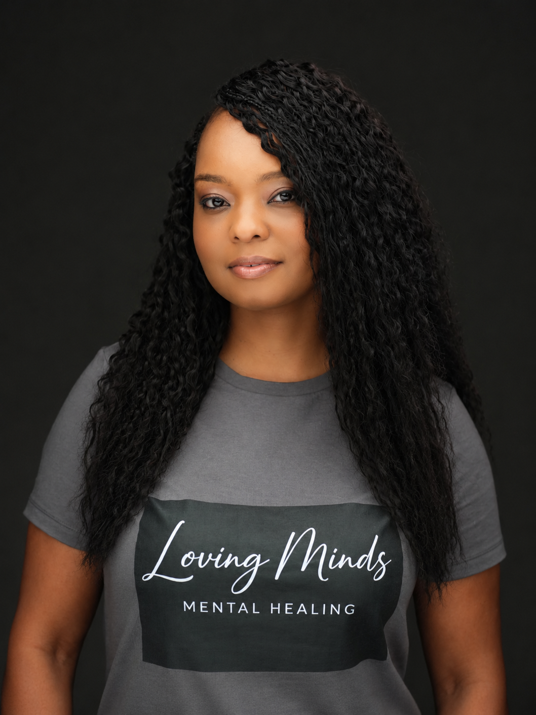 A woman with curly black hair wearing a gray T-shirt with 'Loving Minds Mental Healing' written on it, standing against a dark background.
