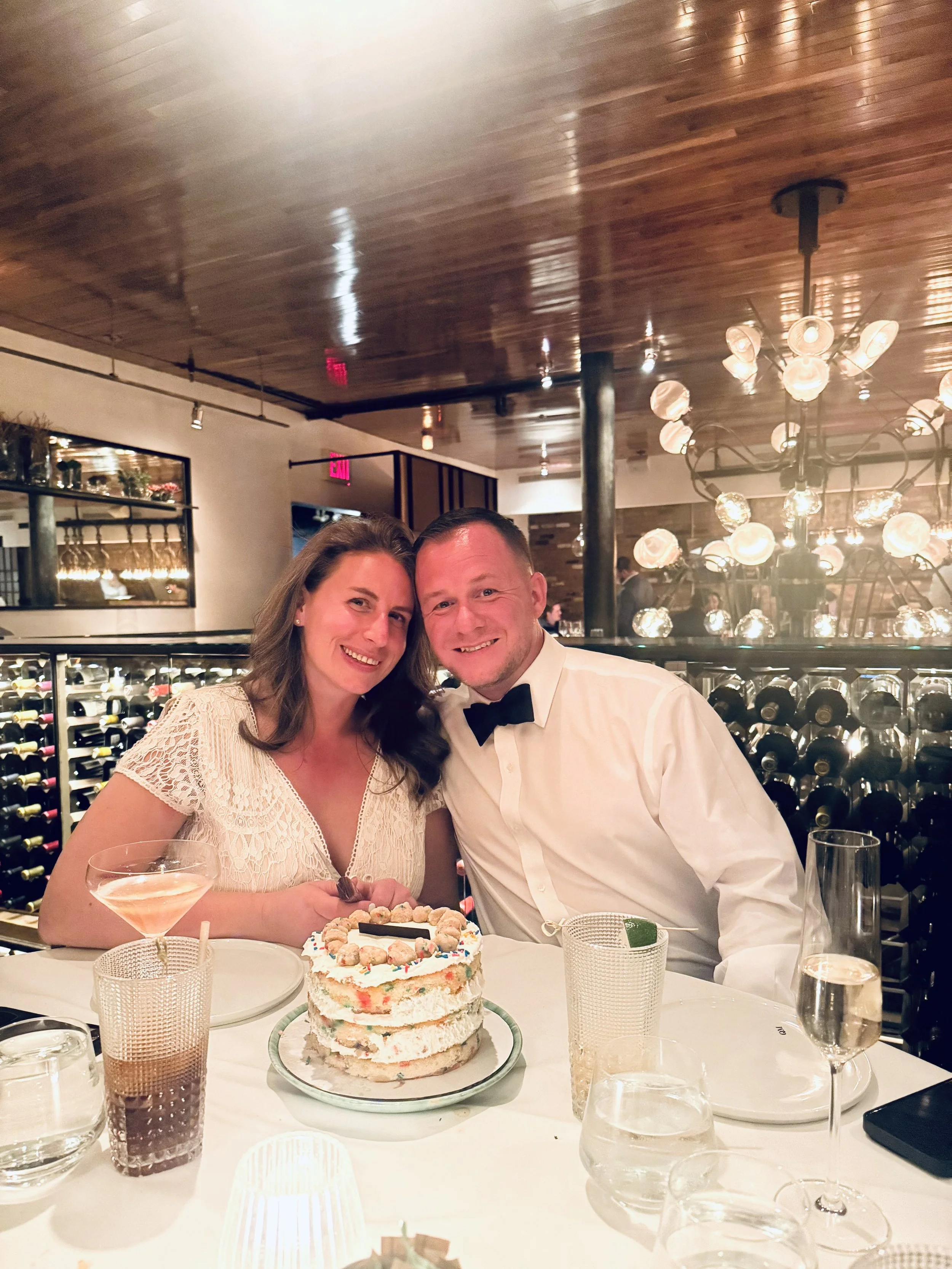 A smiling couple celebrating a birthday in a restaurant, with a layered birthday cake with cookies on top, glasses of drinks, and a wine rack in the background.