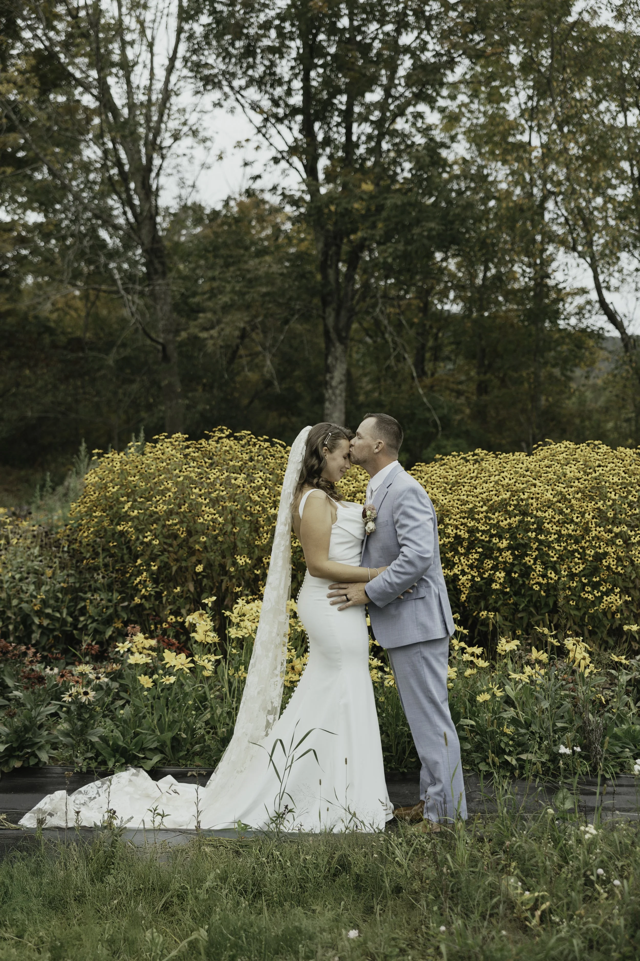 A bride and groom sharing a kiss during their wedding ceremony outdoors in a garden with yellow flowers and trees in the background.