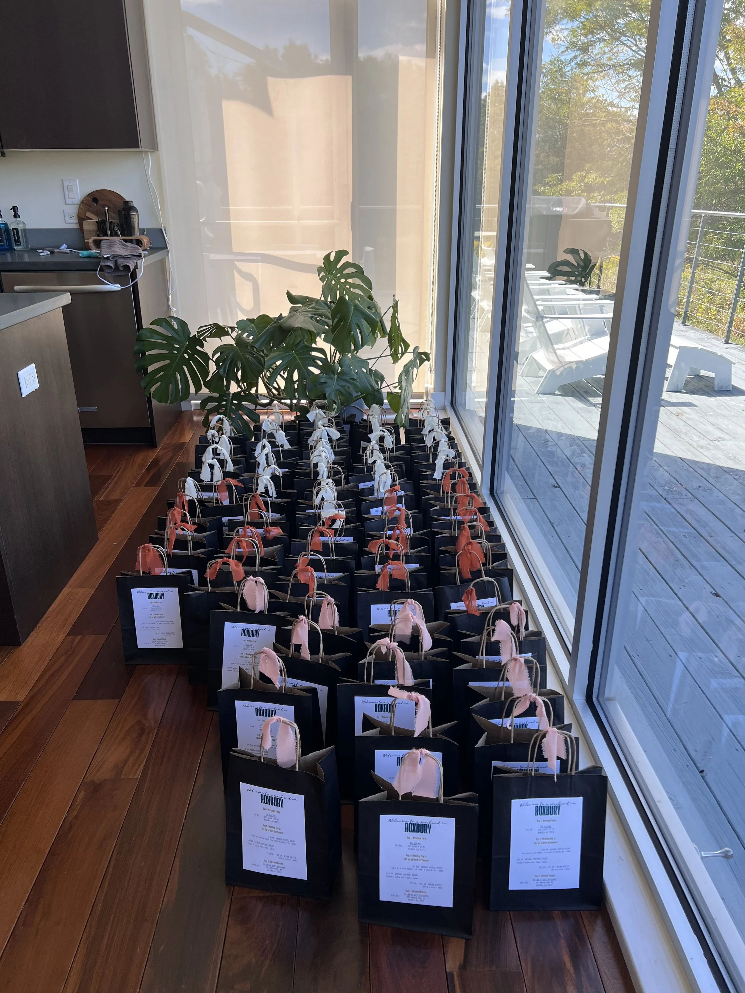 Multiple black gift bags with pink and beige tissue paper, lined up on wooden floor near large glass window, with a potted plant and outdoor patio furniture visible outside.