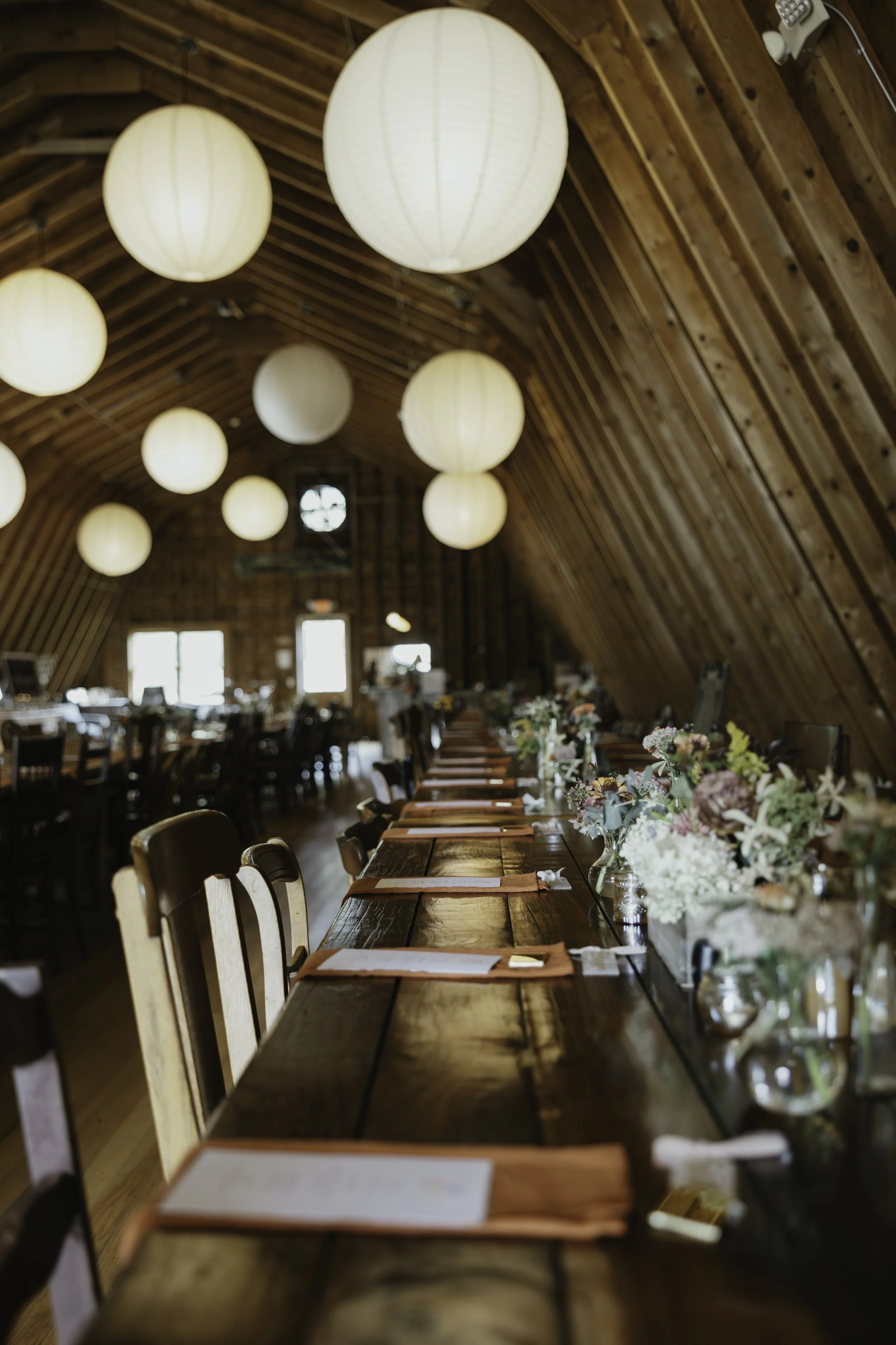 Long wooden dining table with place settings and floral centerpieces inside a rustic, vaulted wooden ceiling restaurant or event space.