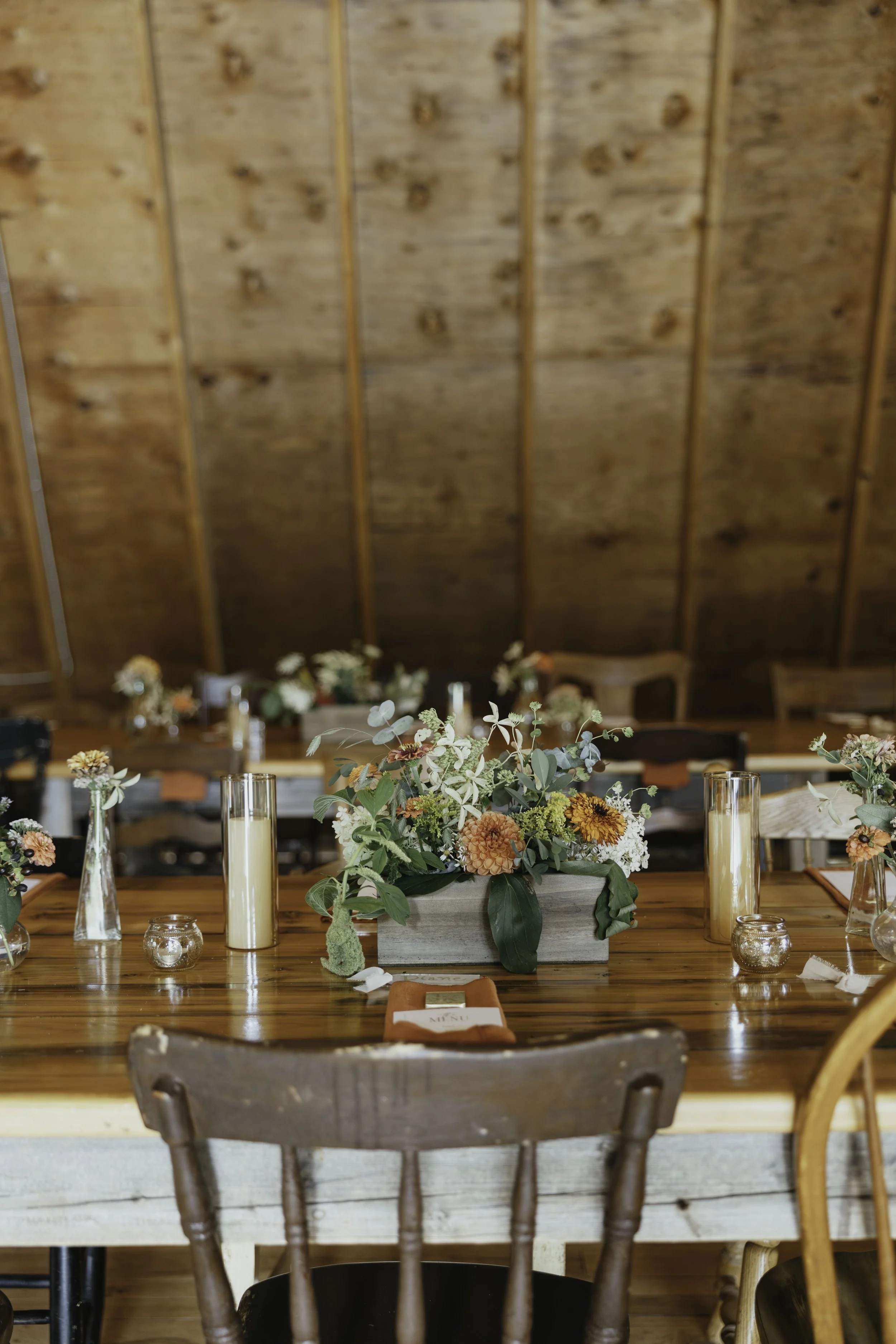 A rustic wooden table decorated with a floral centerpiece, candles, and a menu, set in a cozy room with a wooden ceiling and chairs.