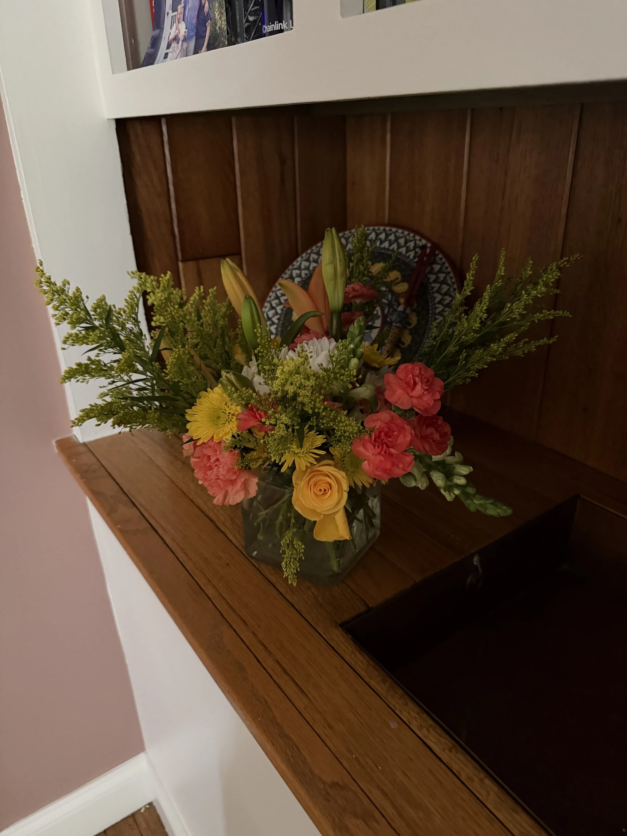 A clear glass vase filled with a colorful assortment of fresh flowers, including yellow roses, pink carnations, orange lilies, white daisies, and greenery, placed on a wooden table against a wall.
