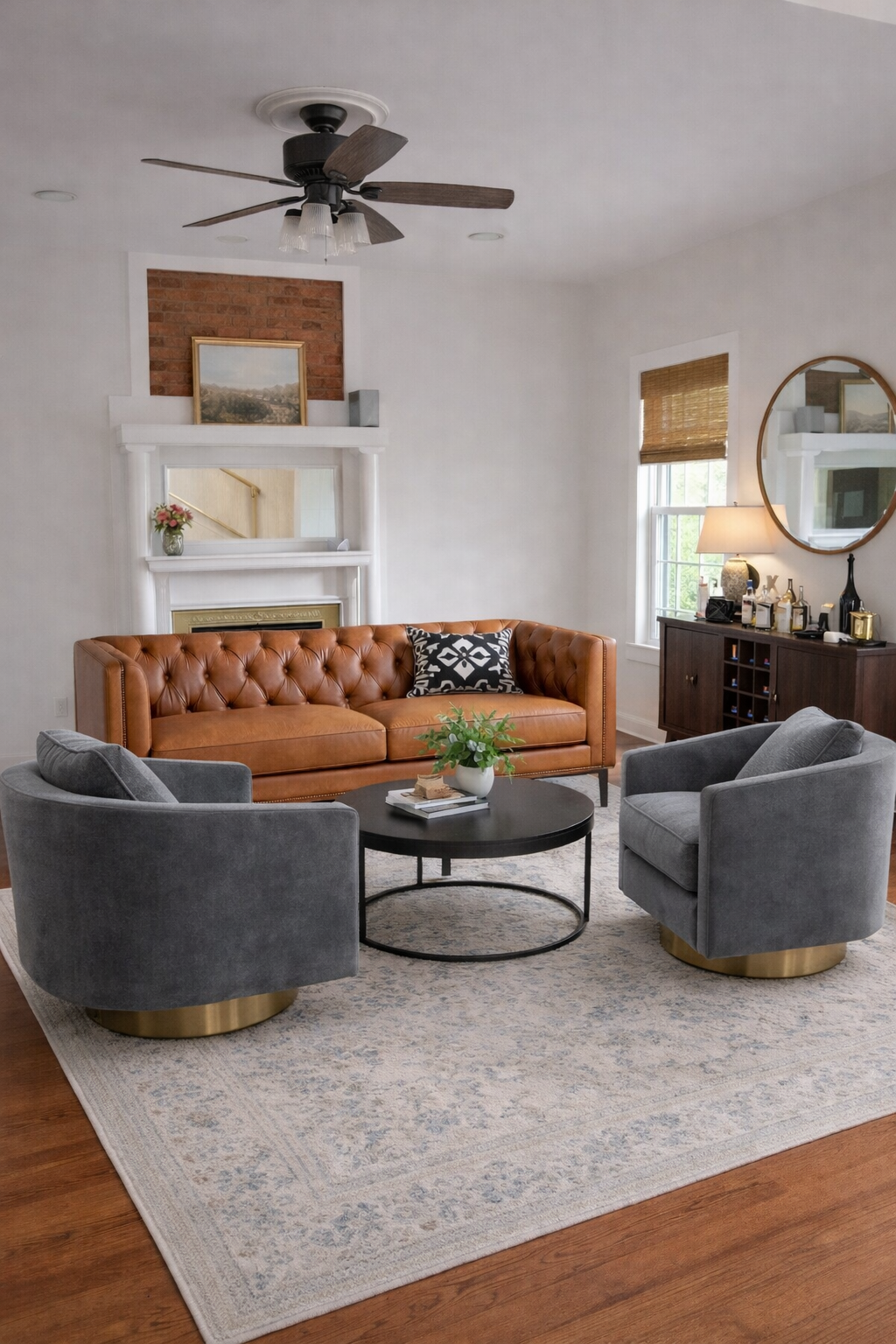 Living room with a brown leather tufted sofa, two gray armchairs, a round black coffee table with a plant, a wooden sideboard, and a white fireplace with a painting above it. There is a ceiling fan and a mirror on the wall, with windows with bamboo b