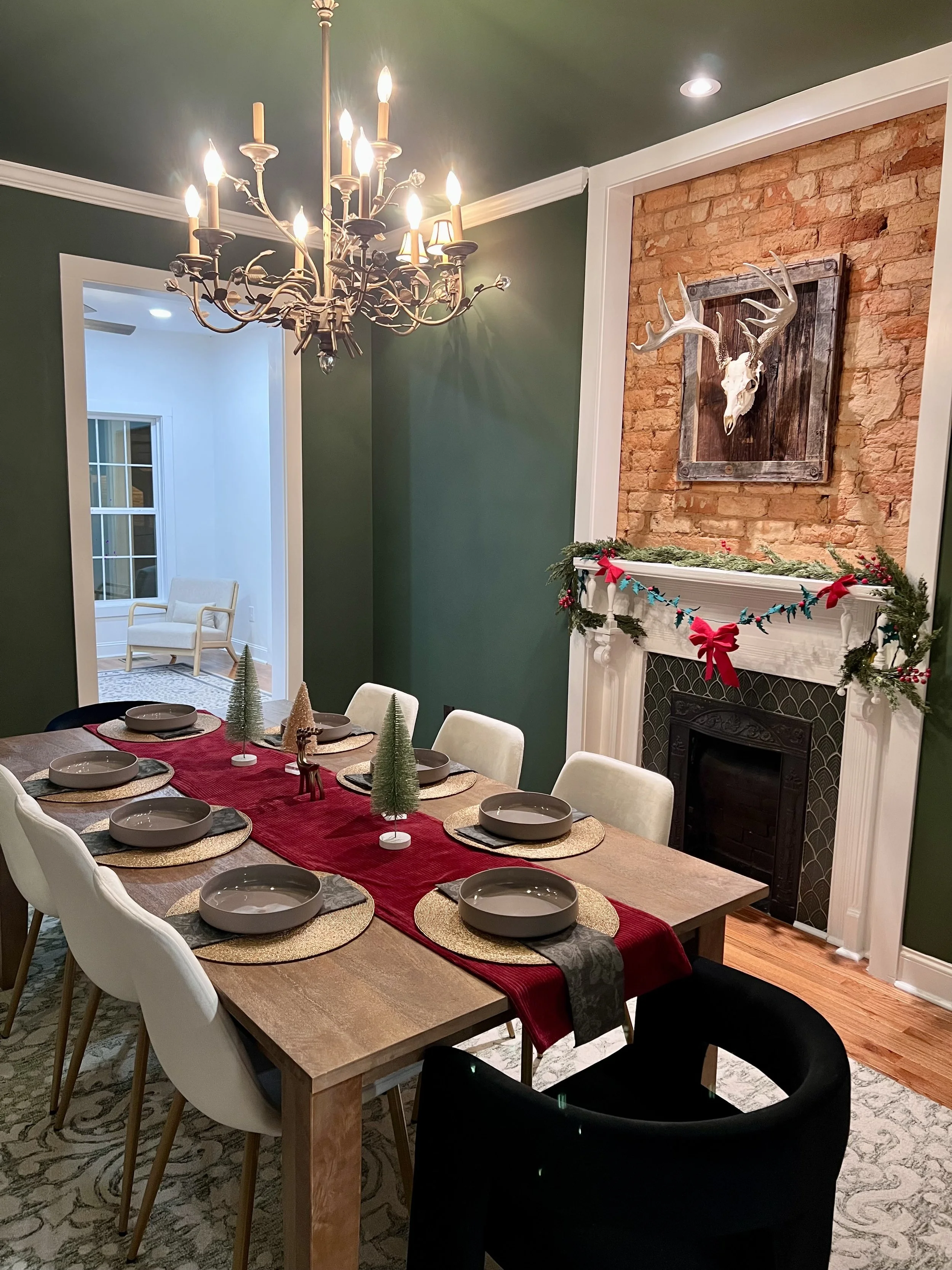 A dining room with a long wooden table set for a holiday meal, decorated with a red table runner, gold placemats, and small Christmas trees, with white chairs around it. There is a fireplace with a garland and red bows, and a brick wall with a mounte
