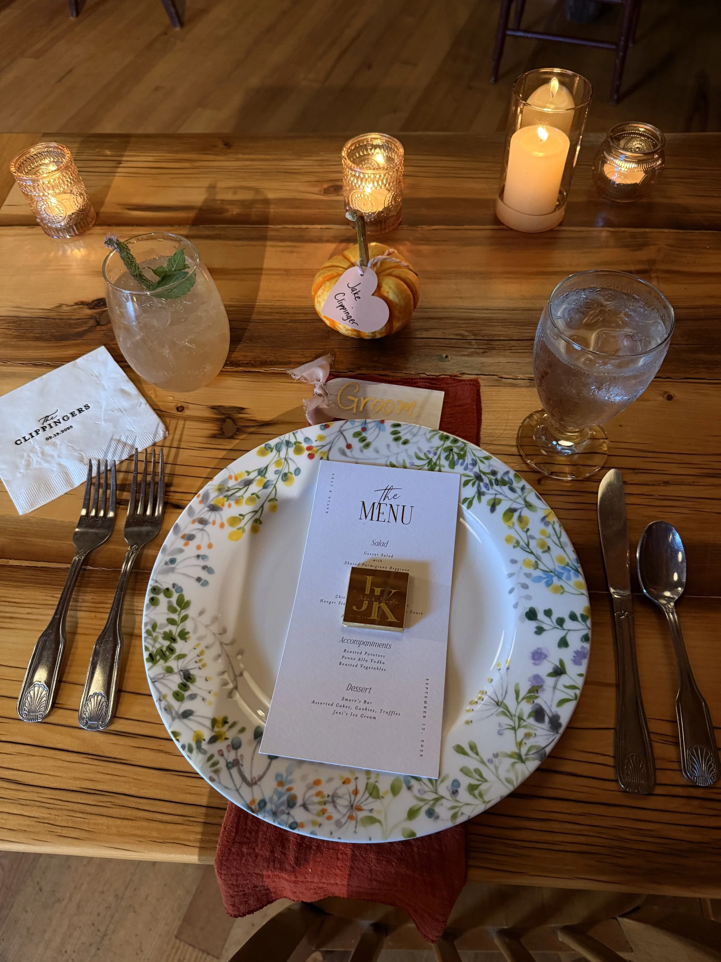 A wedding reception table setup with a floral-patterned plate, a menu card, silverware, candles, and a small pumpkin with a heart-shaped tag reading 'Jake, Cuppinger'. There is a glass of water with ice and a drink garnished with a sprig of mint.