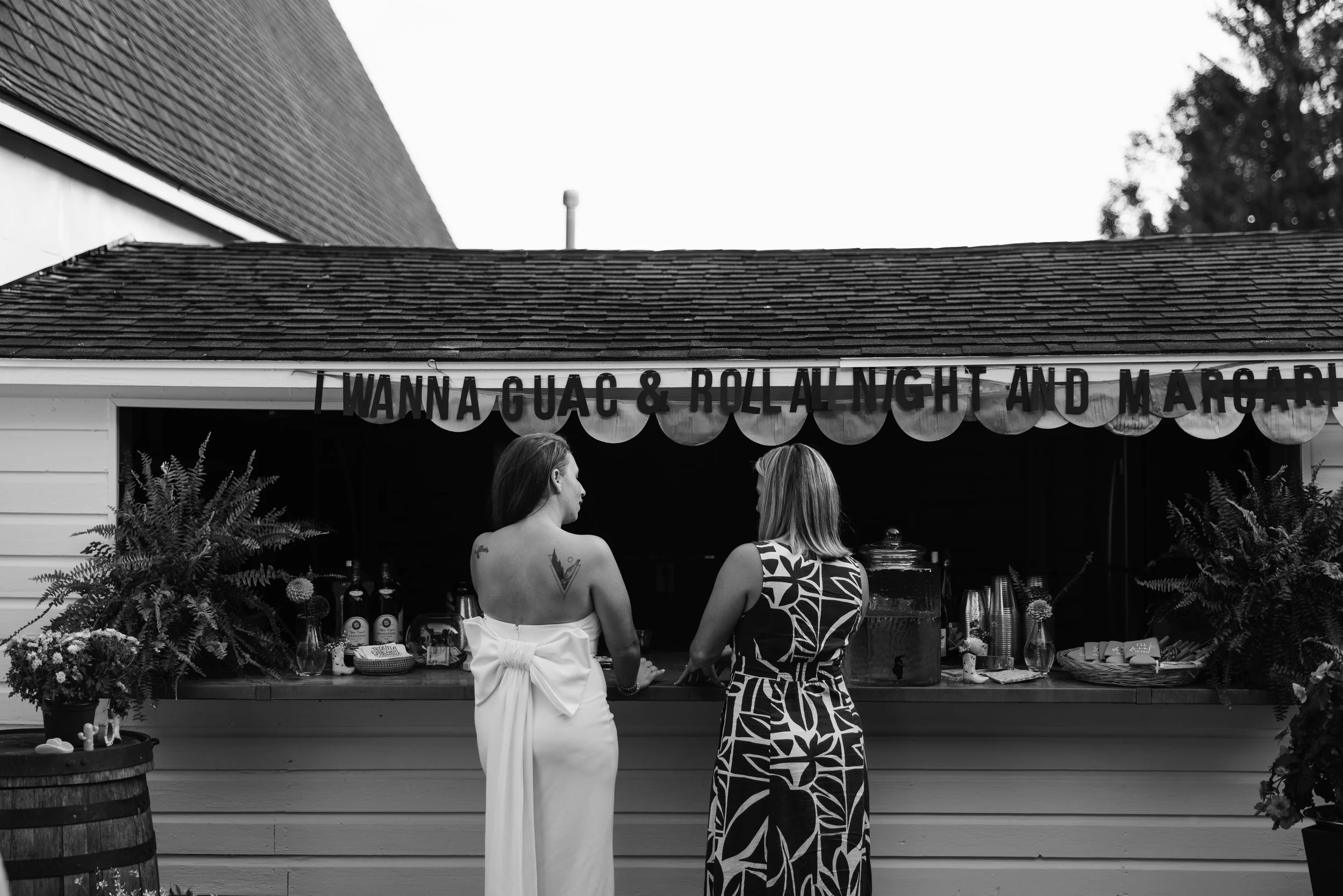 Two women are standing at a food stand with a sign reading 'Wanna Guac & Roll at Night and Margarit.' One woman has tattoos and is wearing a white dress, while the other has shoulder-length hair and is dressed in a patterned dress. The stand has bott