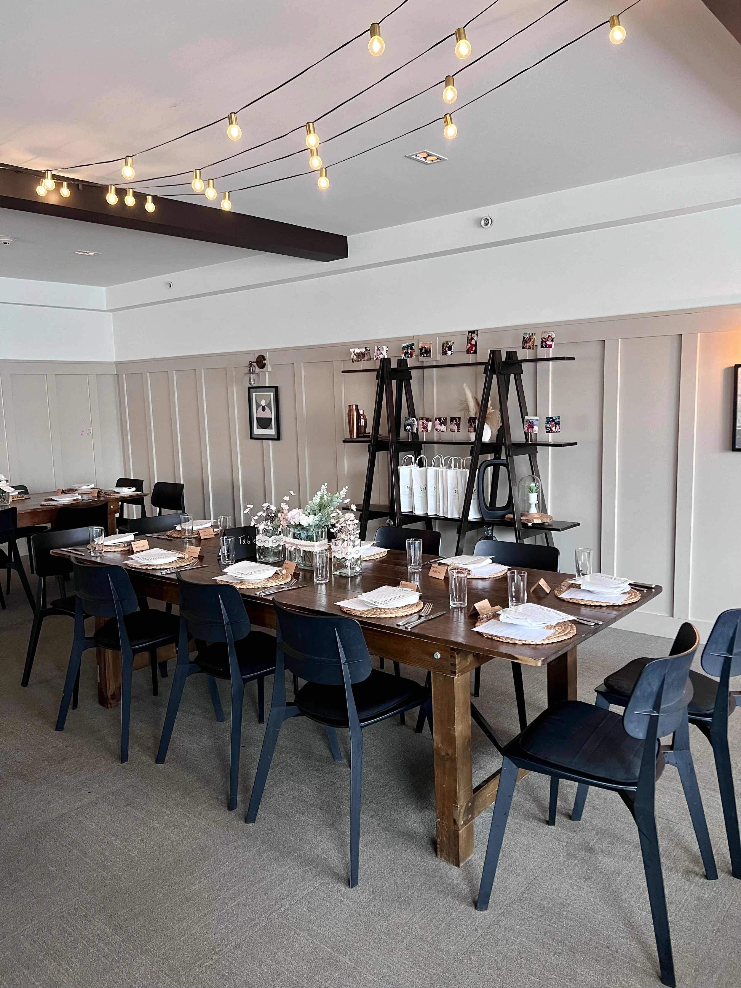 A dining room with a long wooden table set with plates, napkins, glasses, and table numbers. The room has black chairs and a wall with paneling, picture frames, and a black shelf holding photos and decorations. A flower arrangement is on the table, a