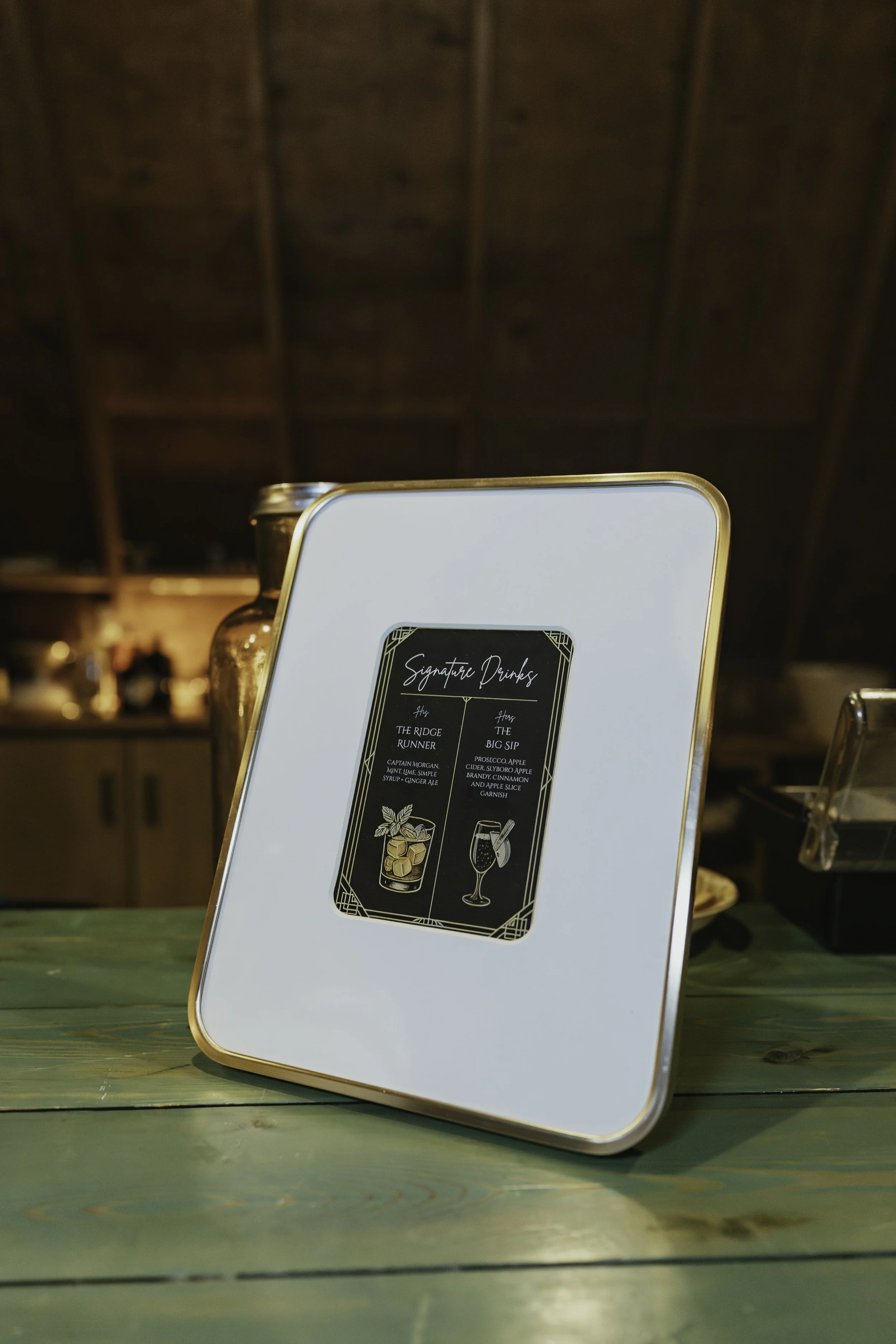 A white framed menu stand on a green wooden table displayed in a dimly lit restaurant or bar setting, with a black and gold sign showing signature drinks.