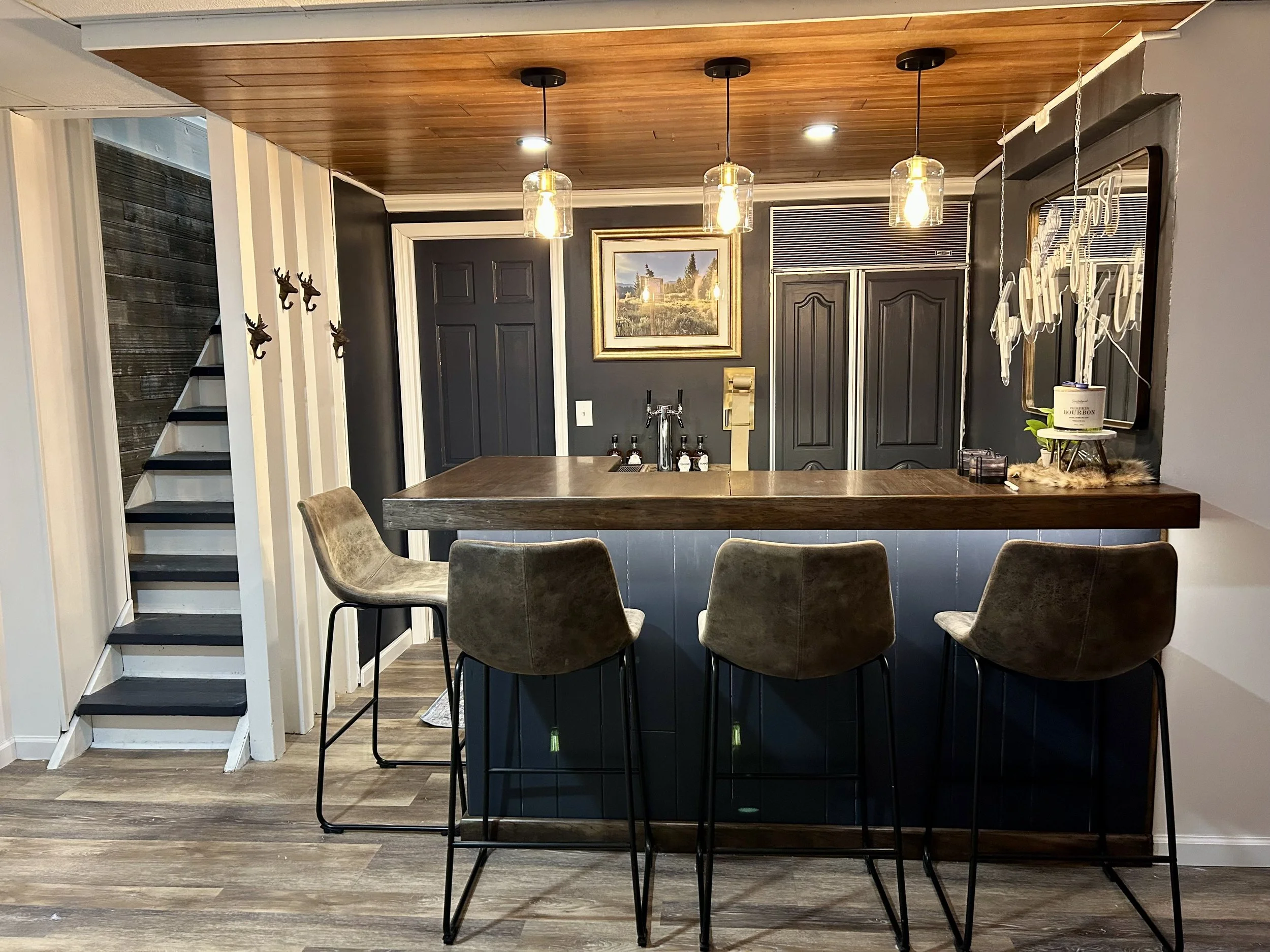 A kitchen island with dark wood panels, three bar stools with cushioned seats, a wall with a framed landscape painting, pendant lights hanging over the island, and decorative elements such as a sign, candles, and a plant.