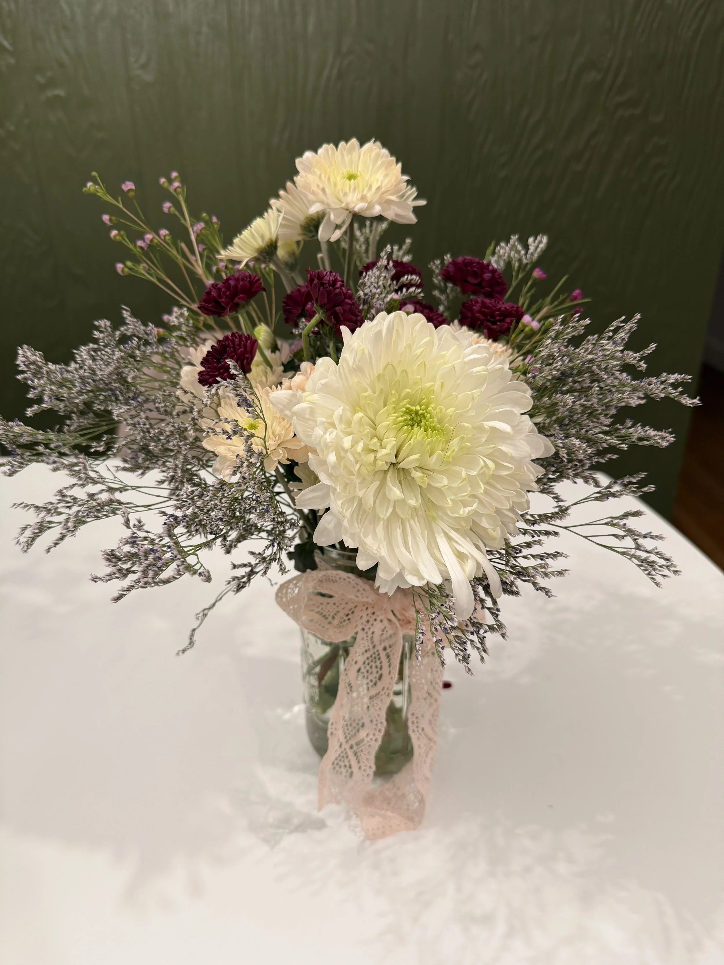 A bouquet of white, purple, and pink flowers with green leaves, wrapped with a pink lace ribbon, in a small glass vase, on a white surface against a green wall.