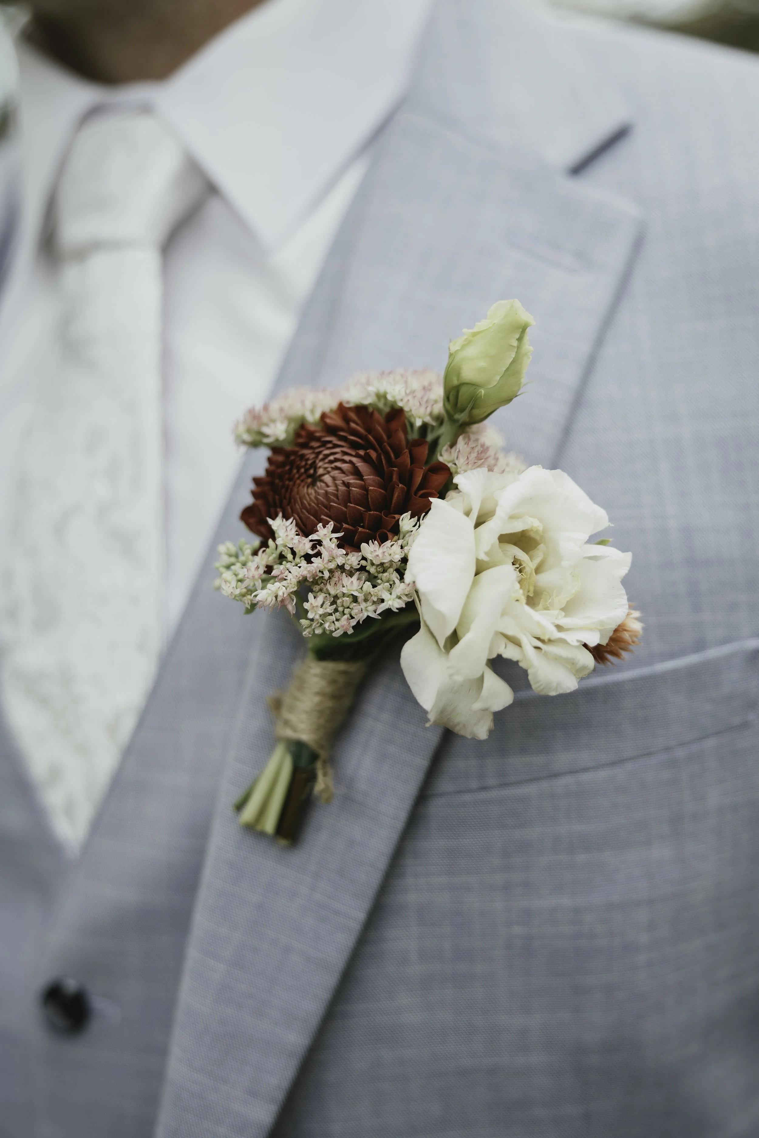A person wearing a light gray suit jacket and a white shirt with a boutonniere featuring white, red, and pink flowers attached to the lapel.