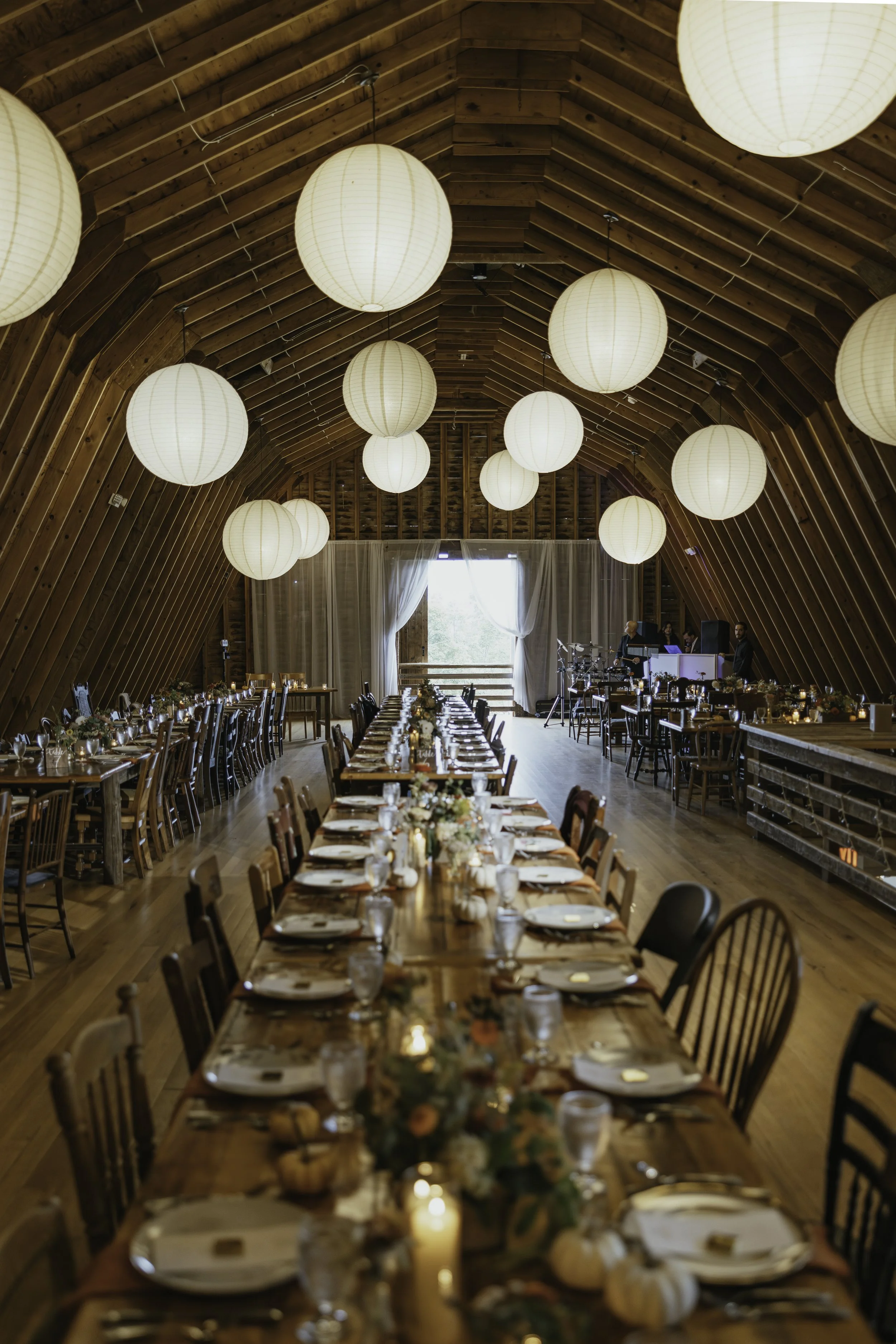 An interior of a rustic wooden barn converted into a banquet hall, decorated with paper lanterns hanging from the ceiling, and set up with long tables decorated for a celebration or event.