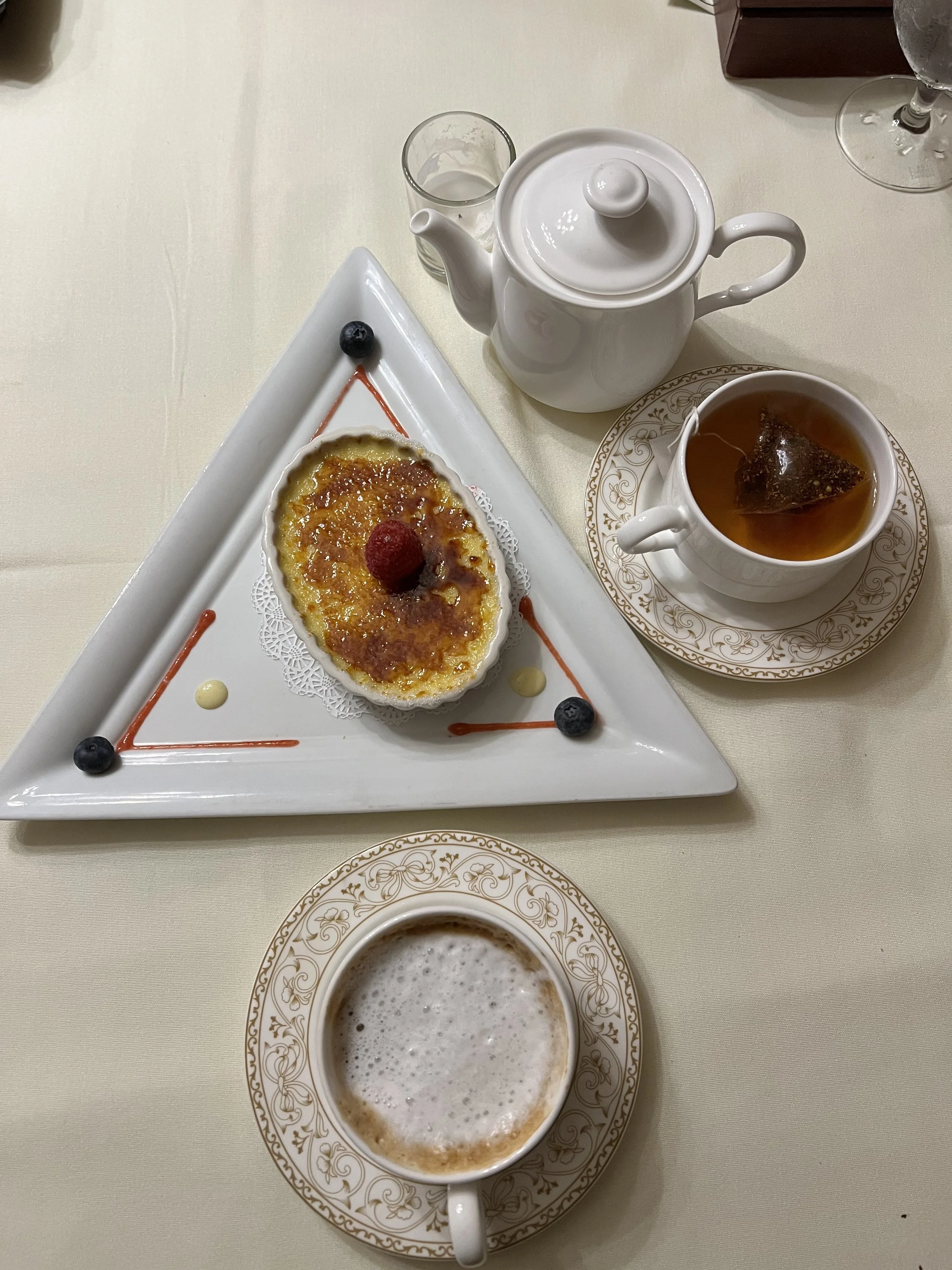 A table with a white teapot, a cup of tea with a tea bag, a glass of water, a cup of a beverage, and a dessert on a white plate. The dessert looks like a flan garnished with berries and sauce.
