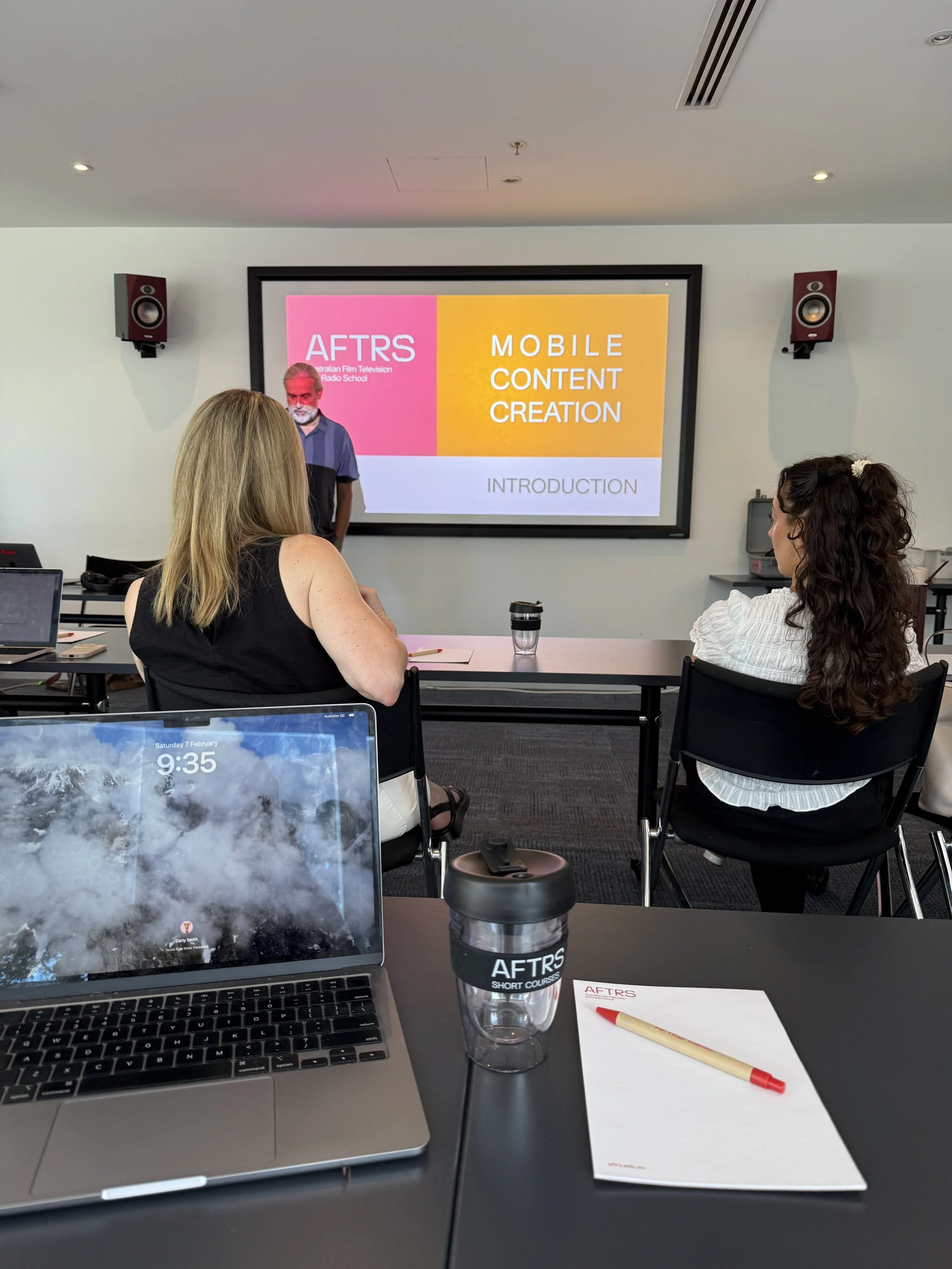 Conference room with a presentation slide on the screen titled 'AFTRS' and 'Mobile Content Creation'. There are two women seated at a table, one with blonde hair and a sleeveless black top, and the other with dark curly hair and a white blouse. A man
