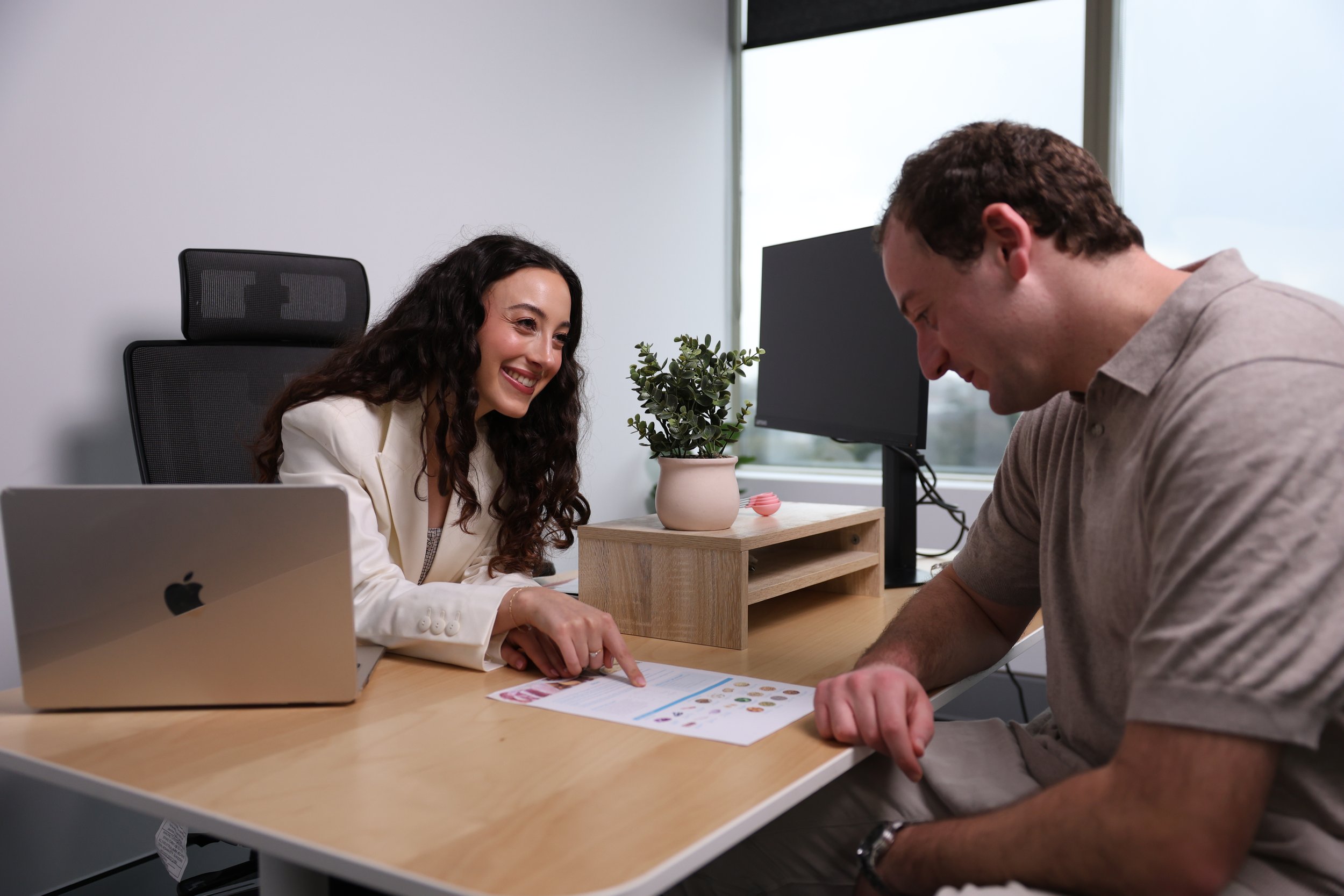 A woman and a man are sitting at a desk, smiling and looking at documents and a printed infographic with icons and graphs, in a modern office with a computer monitor and a potted plant.