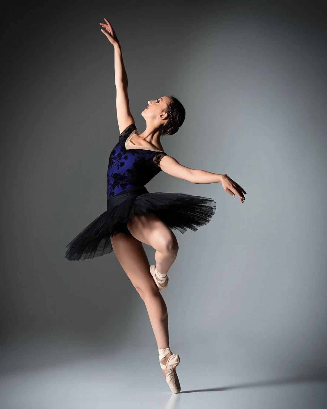 A ballerina in a black and blue tutu performing a pose on pointe against a gray background.