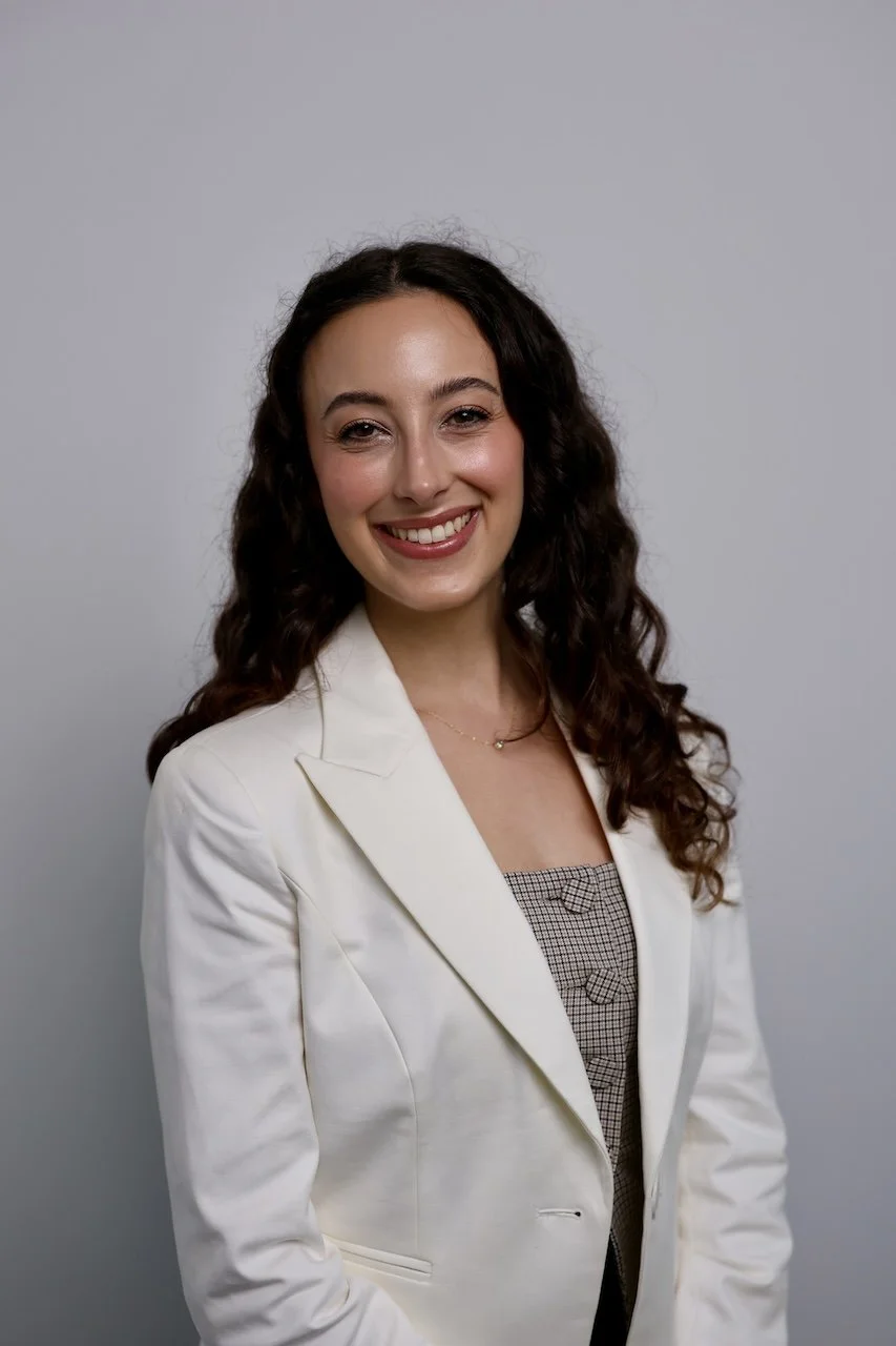 A woman with long curly brown hair smiling, wearing a white blazer over a patterned top, against a light gray background.