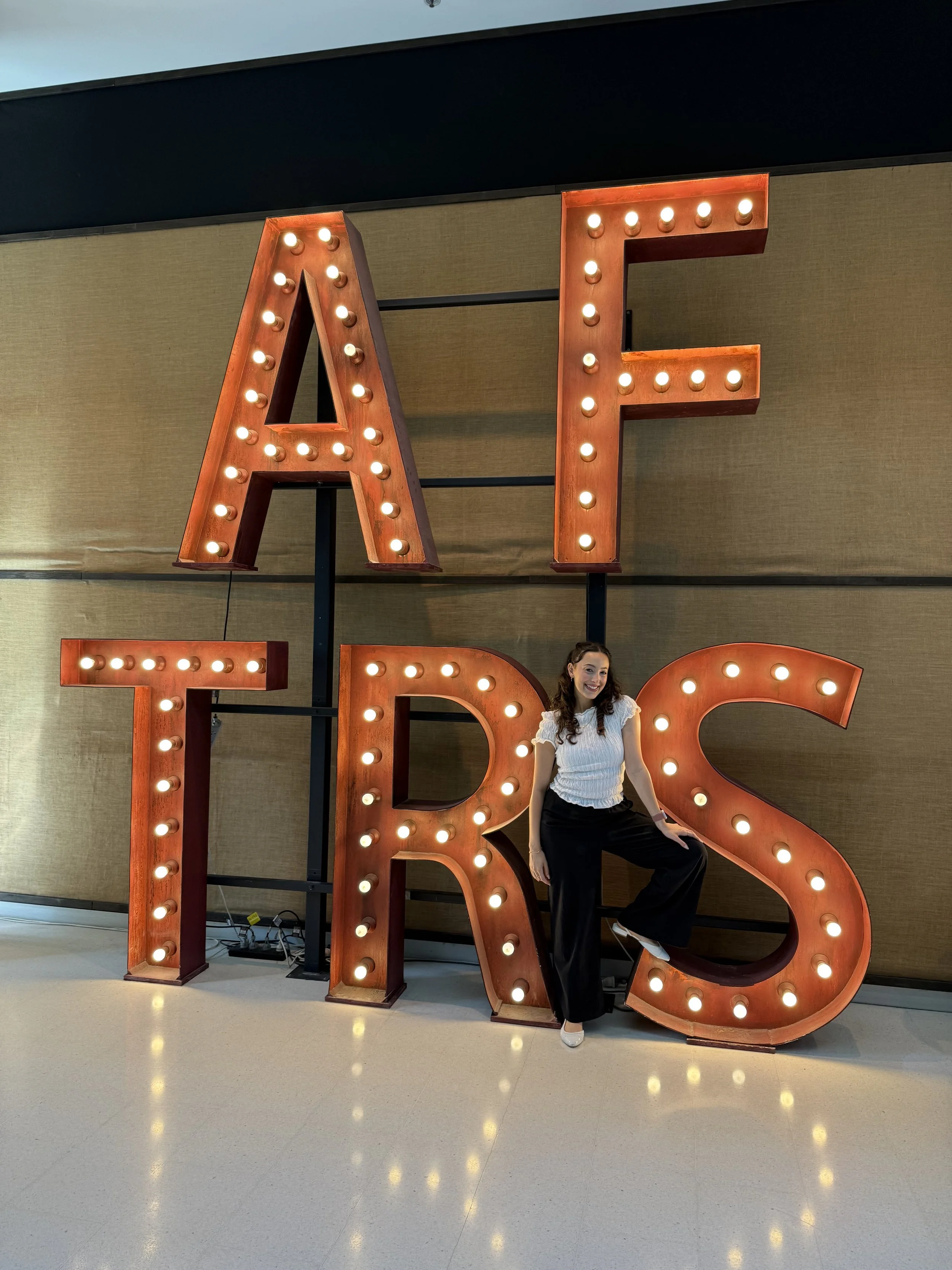 A woman with dark hair and a white blouse sitting on a large vintage-style marquee letter 'R' with lights, positioned next to illuminated birthday letters spelling 'BIRTHDAY'.