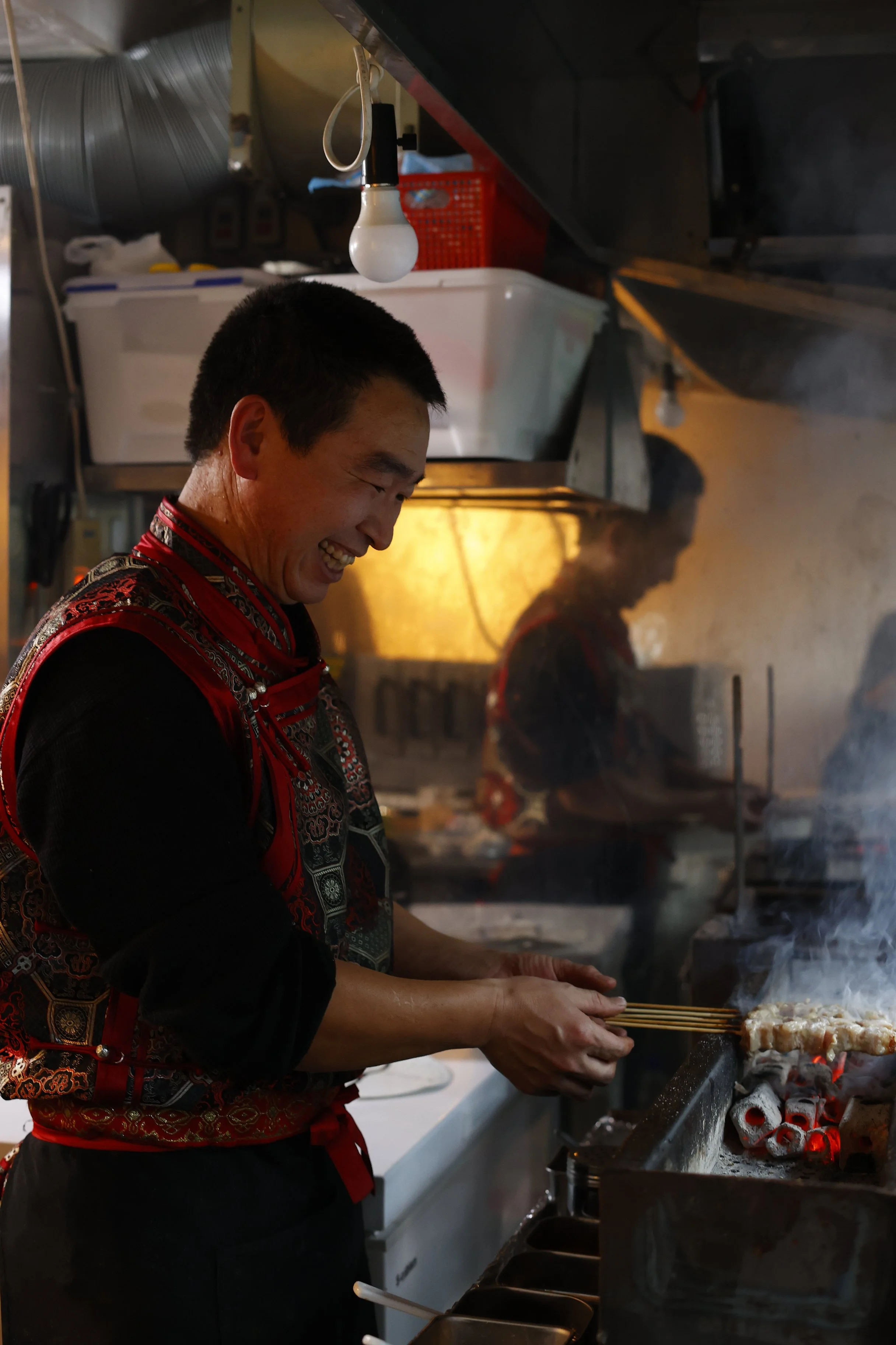 A man grilling skewered food over charcoal on a grill in a kitchen, smiling as he cooks.