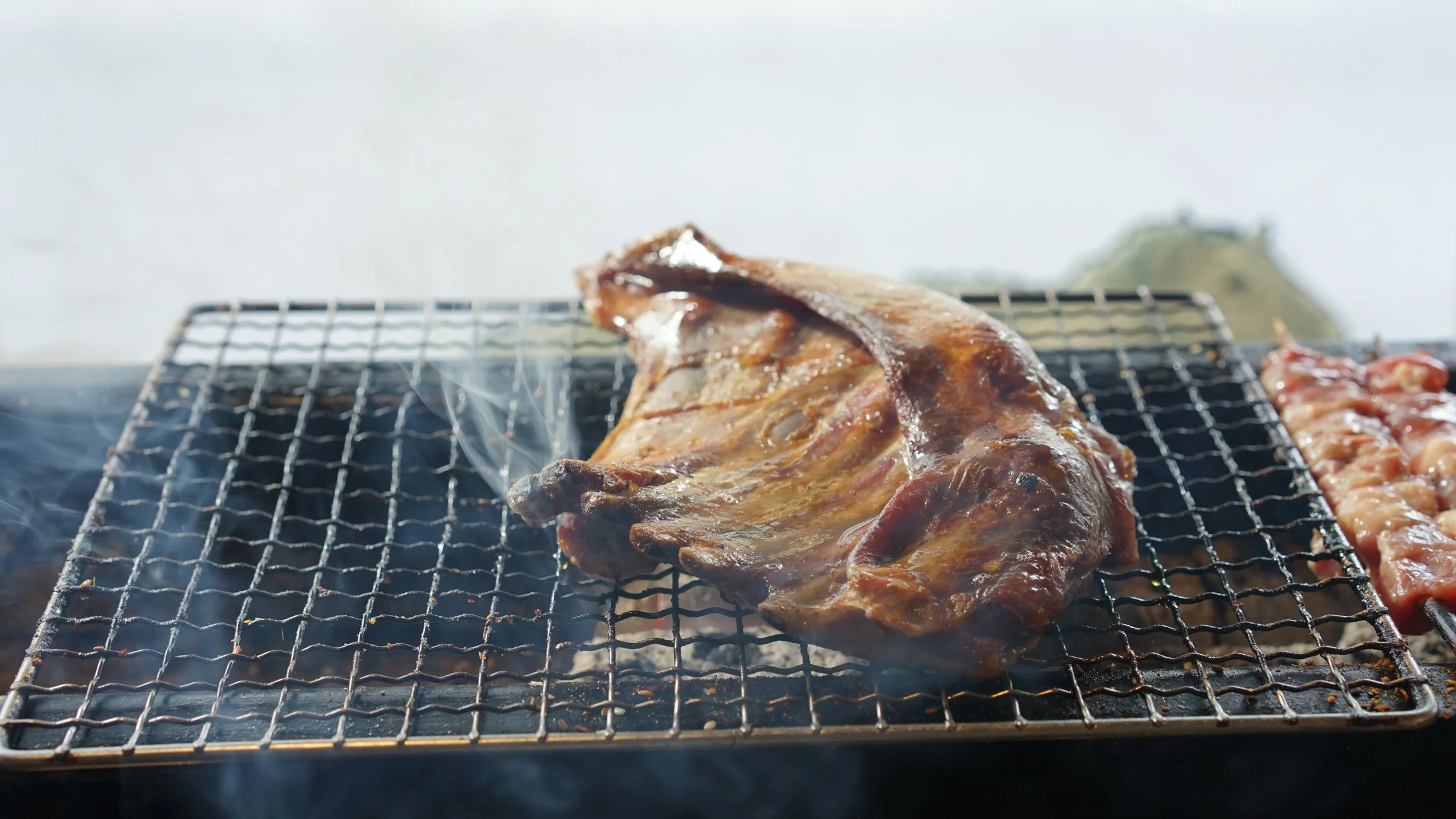 Close-up of a cooked steak on a grill with smoke rising, with some meat pieces visible on the side.