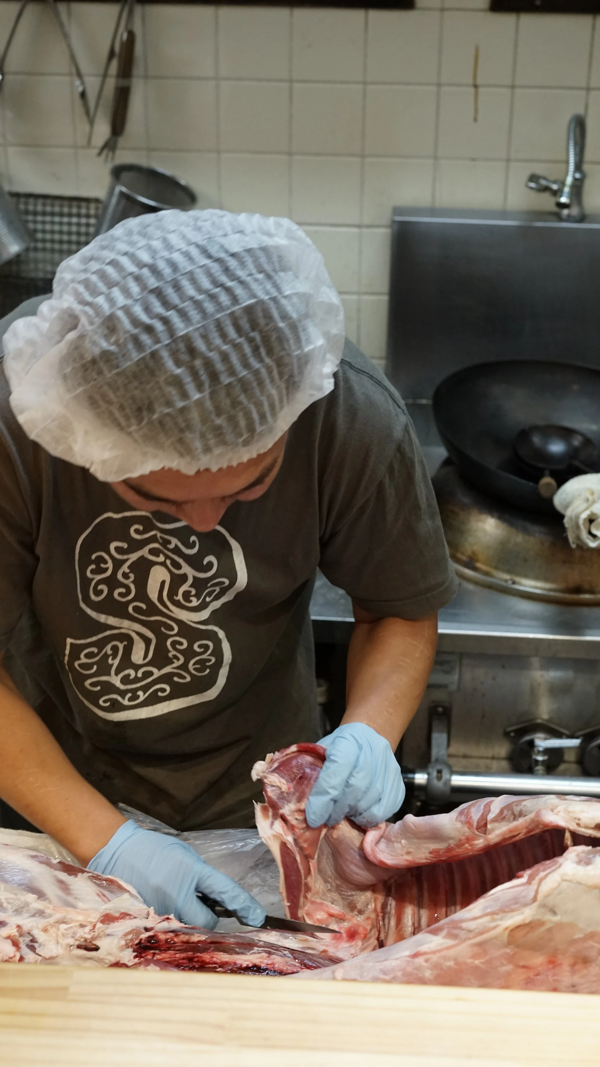A butcher wearing a hairnet and gloves is cutting meat on a worktable in a kitchen. There are kitchen tools and a stove in the background.
