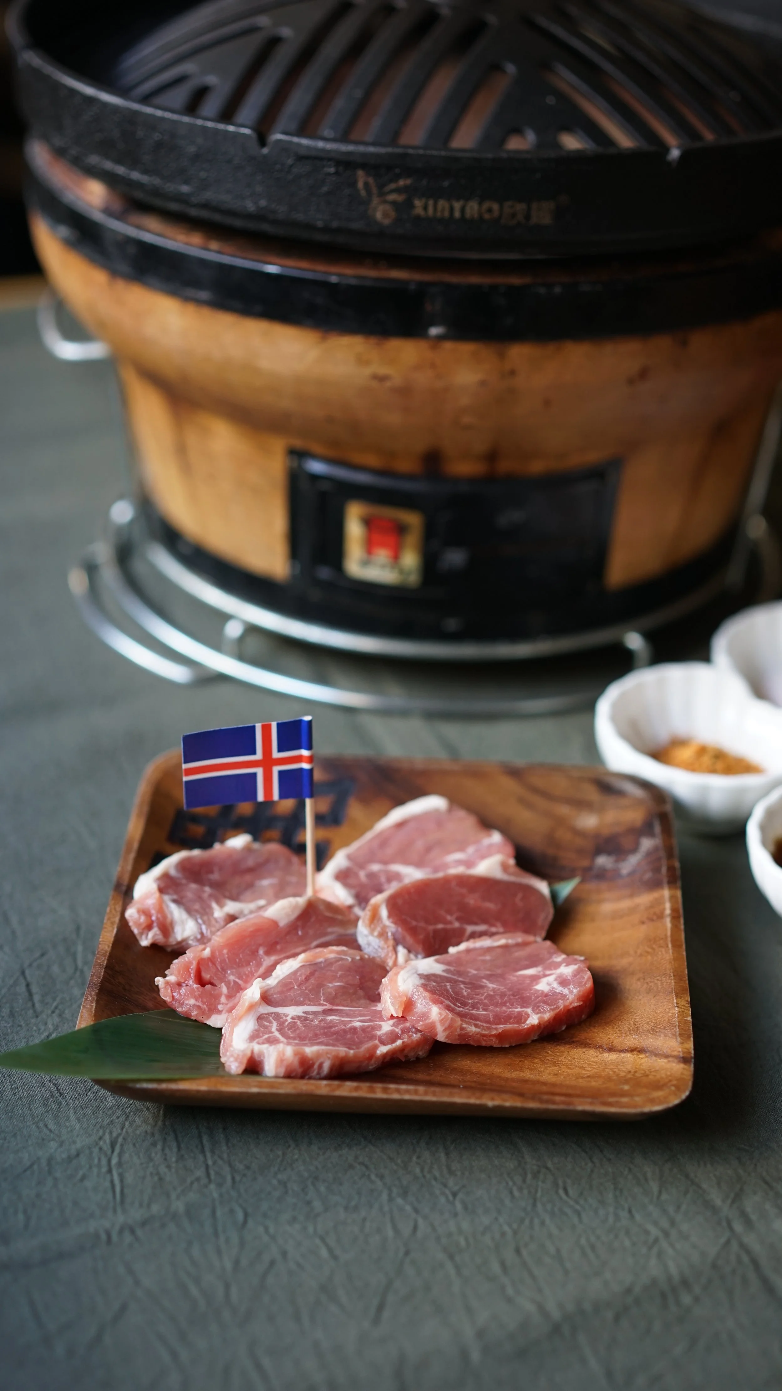 Raw pork slices on a wooden tray with a small Icelandic flag, alongside bowls of seasonings, in front of a tabletop rotisserie grill.