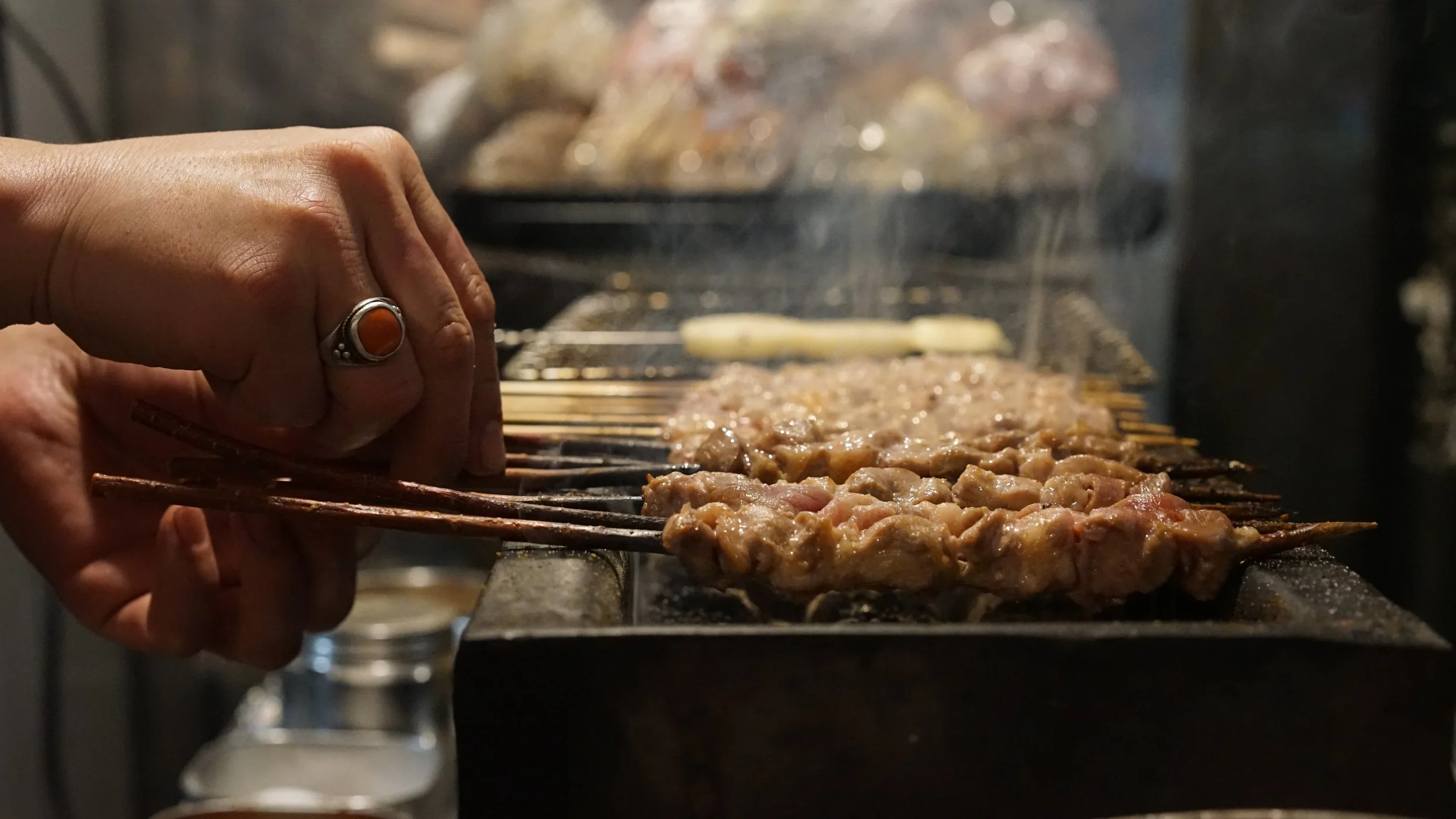 Person grilling skewers of meat over an open flame.