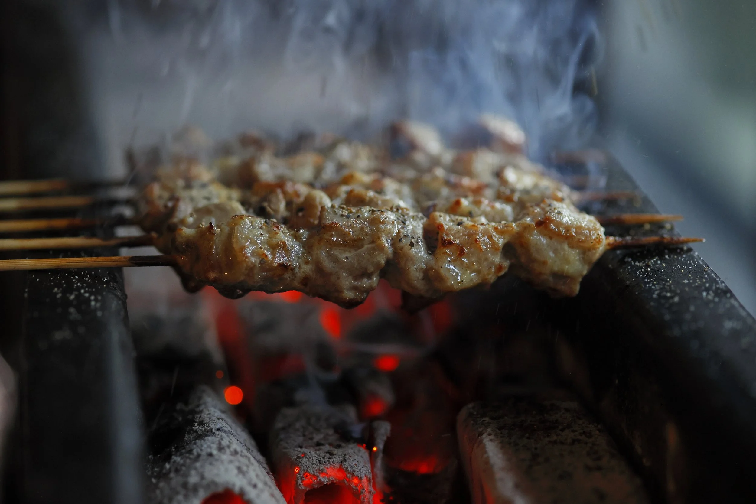 Skewers of meat cooking over hot coals with smoke rising, on a barbecue grill.