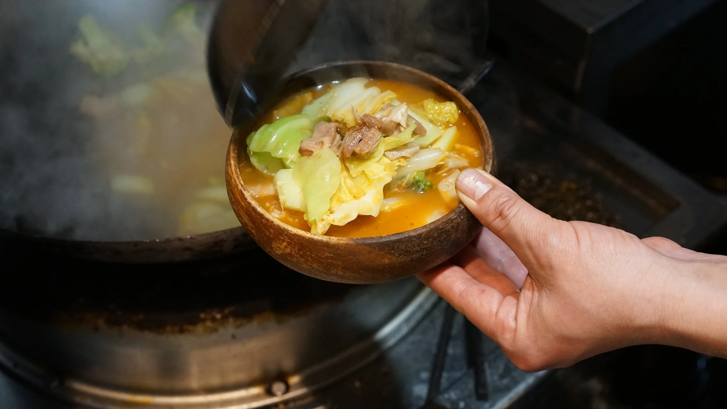 Person holding a wooden bowl of hot soup with vegetables and meat over a stove.